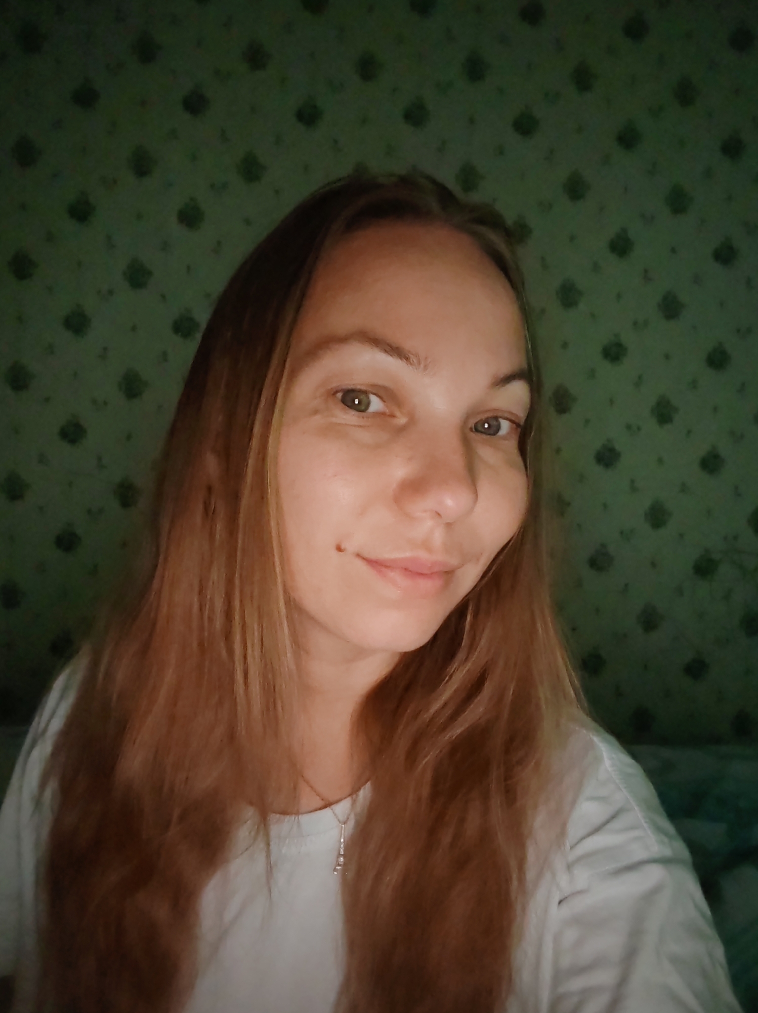 A young woman with long, light brown hair and a mole on her cheek is pictured in a close-up, head-and-shoulders shot. She is wearing a white t-shirt and a delicate necklace. The background is a green, patterned wallpaper. The lighting is dim, suggesting an indoor setting. There is no indication of the specific location within the city of Чухлэм, Russia.