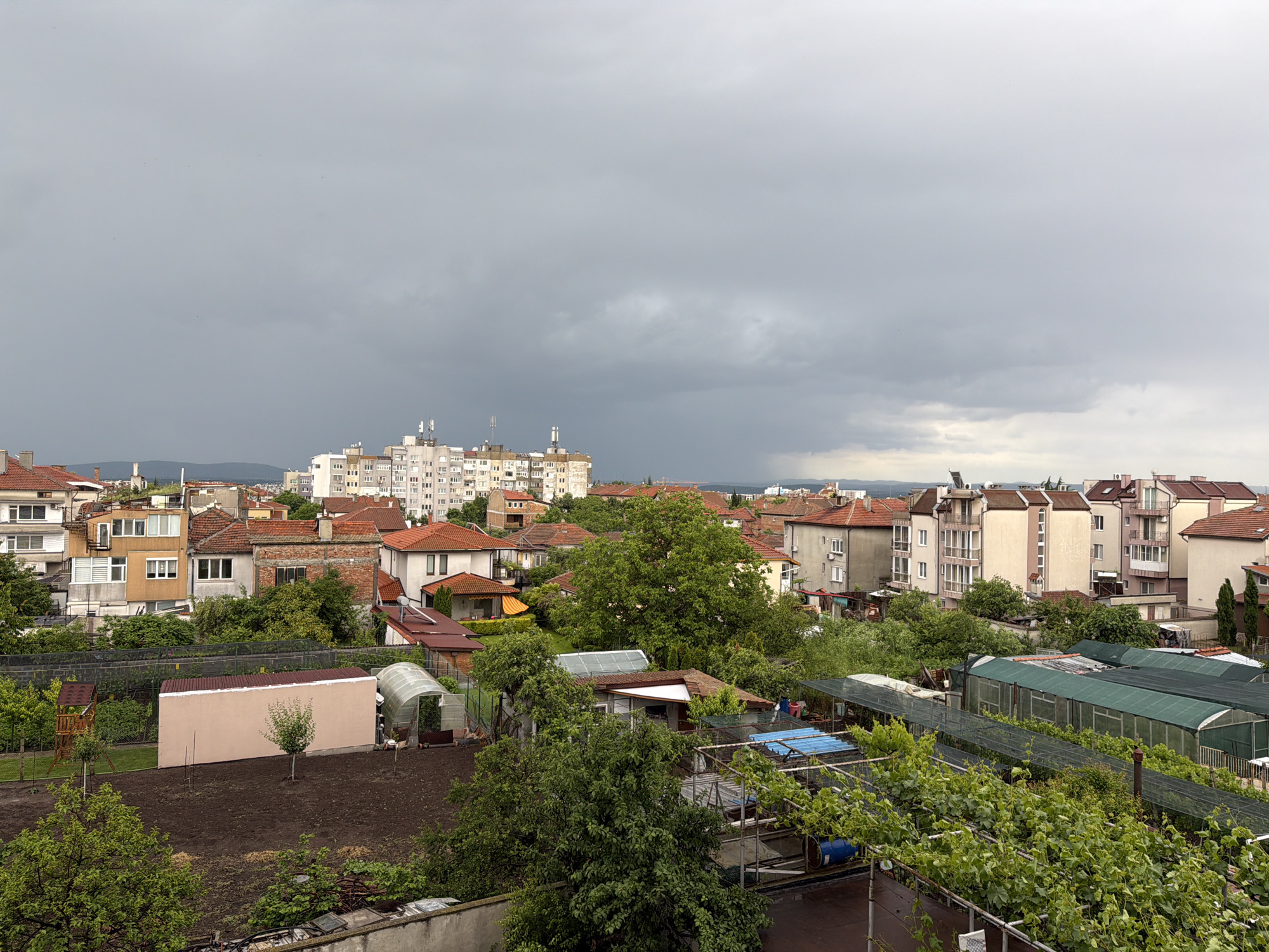 A medium, eye-level shot shows a rooftop perspective of the city of Burgas, Bulgaria. The sky is overcast with dark, low-hanging storm clouds. The cityscape is a mix of residential buildings with red-tiled roofs, interspersed with green trees and garden plots. In the foreground, there are multiple greenhouses with green coverings and various garden structures, including a trellis with vines. A tan-colored shed stands in a plowed garden plot. In the midground, various houses with red tile roofs and apartment buildings can be seen. In the distance, more apartment buildings are visible on the horizon.
