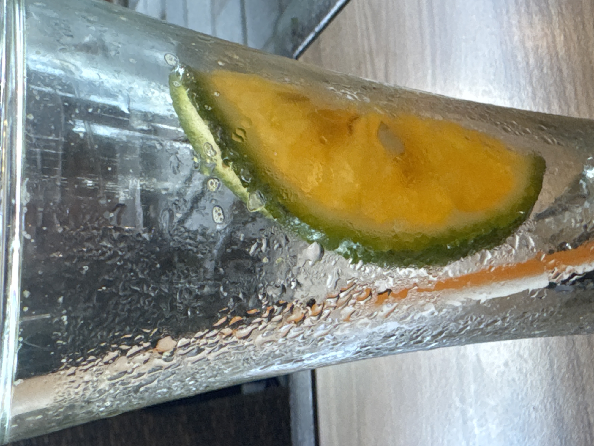A close-up, eye-level shot captures a glass of iced beverage, likely in a city like Nha Trang, Vietnam, given the presence of a beverage commonly served in such locations. The glass is filled with a clear liquid and contains a slice of citrus fruit, possibly lime or orange, with its rind visible. The exterior of the glass is covered in condensation, indicating it is chilled. Bubbles are visible within the liquid, suggesting it is carbonated. An orange-colored straw is partially submerged in the drink. The background is out of focus, showing hints of outdoor seating and a muted, diffused light. The scene suggests a casual setting, perhaps a cafe or restaurant on a warm day.