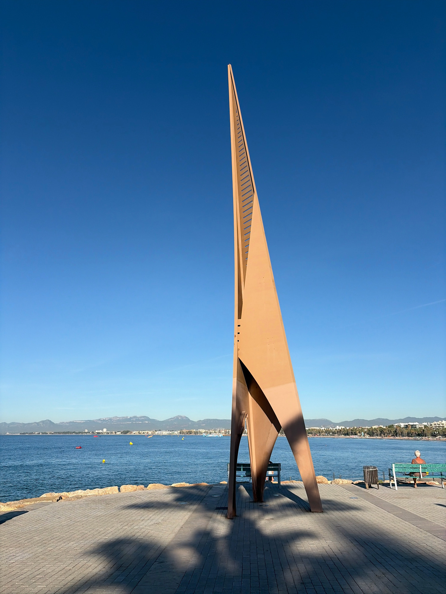 The image shows a tall, abstract sculpture made of tan-colored metal, positioned on a paved walkway next to the sea. The sculpture has a triangular, tapering form with a series of horizontal openings along one edge, resembling a ladder or a sail. Its shadow is cast across the walkway in the foreground. To the right of the sculpture, two park benches are visible. On the farther bench, a person with white hair is seated, facing away from the camera and towards the water. A trash can is located between the two benches.
The walkway is paved with gray rectangular tiles and bordered by a low stone wall separating it from the blue sea. In the background, across the water, is a coastline with buildings, palm trees, and mountains in the distance under a clear, bright blue sky. The scene appears to be a public waterfront area in the city of Salou, Spain, likely a promenade or esplanade. The lighting suggests it is daytime, with the sun casting distinct shadows from the sculpture.