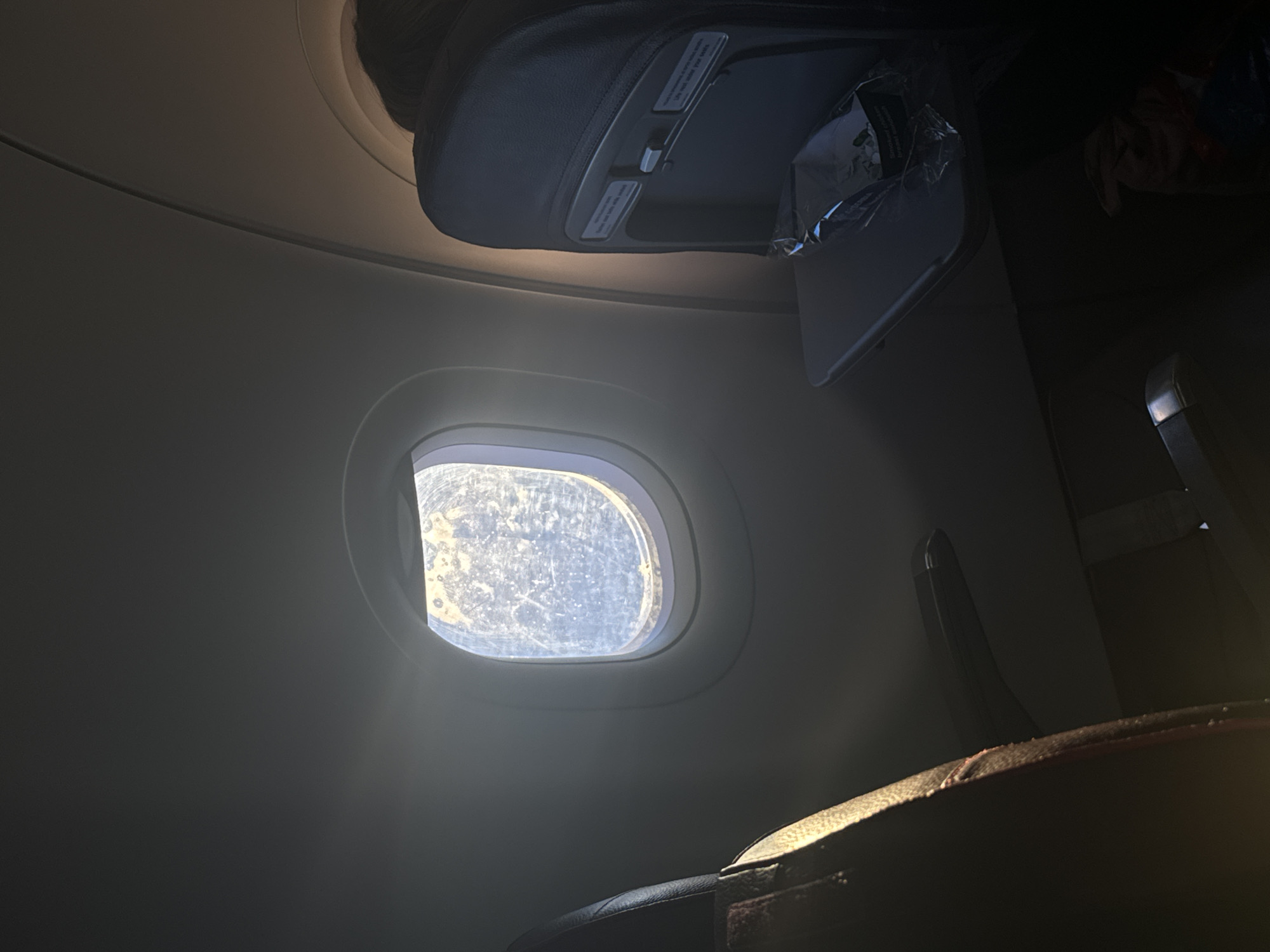 The image depicts the interior of an airplane cabin, with a focus on a window and its surroundings. Sunlight streams through the window, which is slightly dirty. Above the window, an overhead compartment is visible, along with a folded-down tray table. A portion of a seat is seen in the foreground. The location, city of Moscow, Russia is not provided within the image.