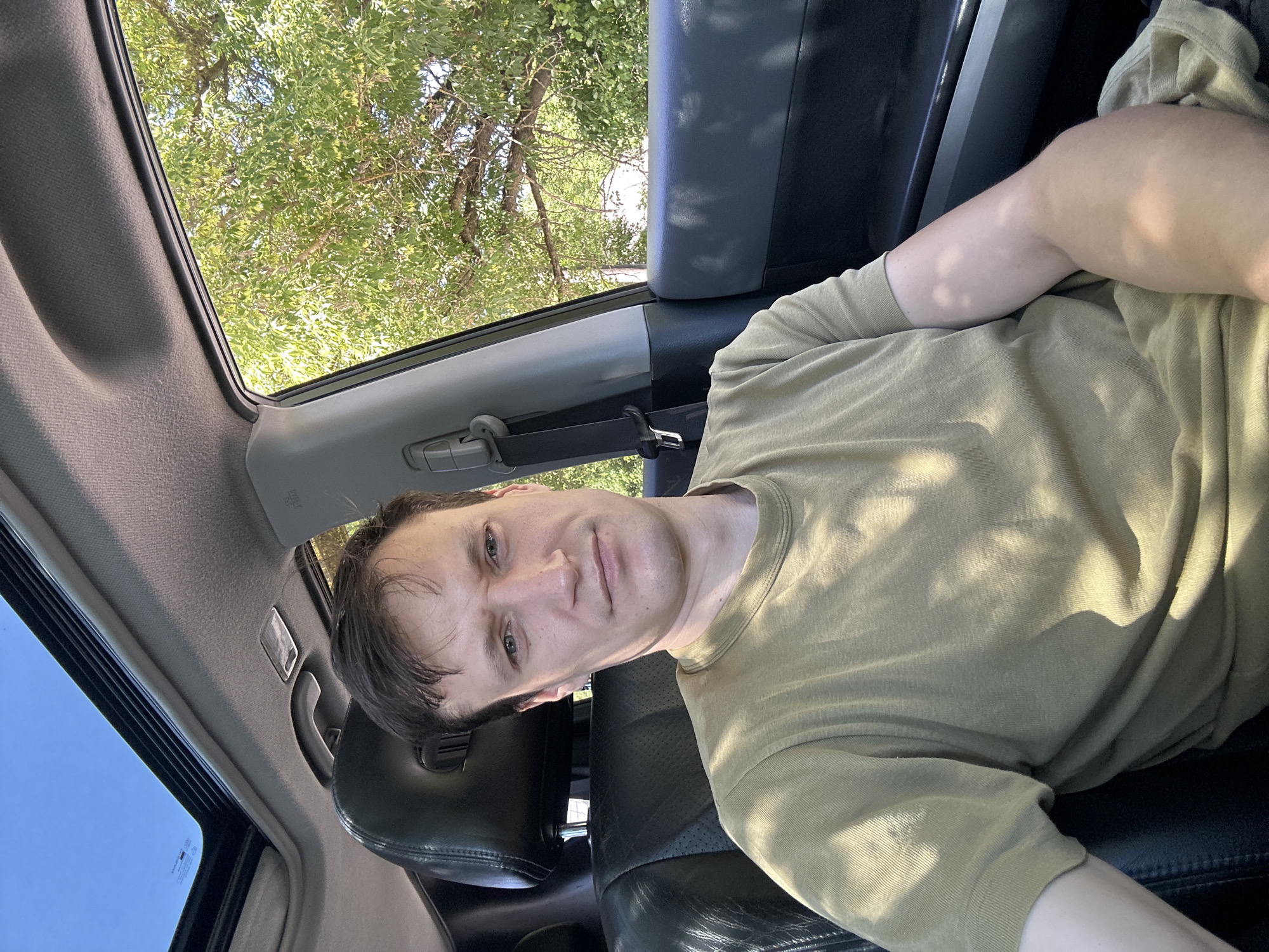 A man with short, dark brown hair and light skin is sitting in the driver's seat of a car, looking directly at the camera. He is wearing a tan t-shirt. The car's sunroof is open, revealing a bright blue sky. Sunlight filters through the leaves of a large tree outside the car window, casting dappled shadows on the man and his shirt. The interior of the car is visible, including black leather seats and a gray headliner. The image appears to have been taken during the daytime. The location is Makiv, Ukraine.