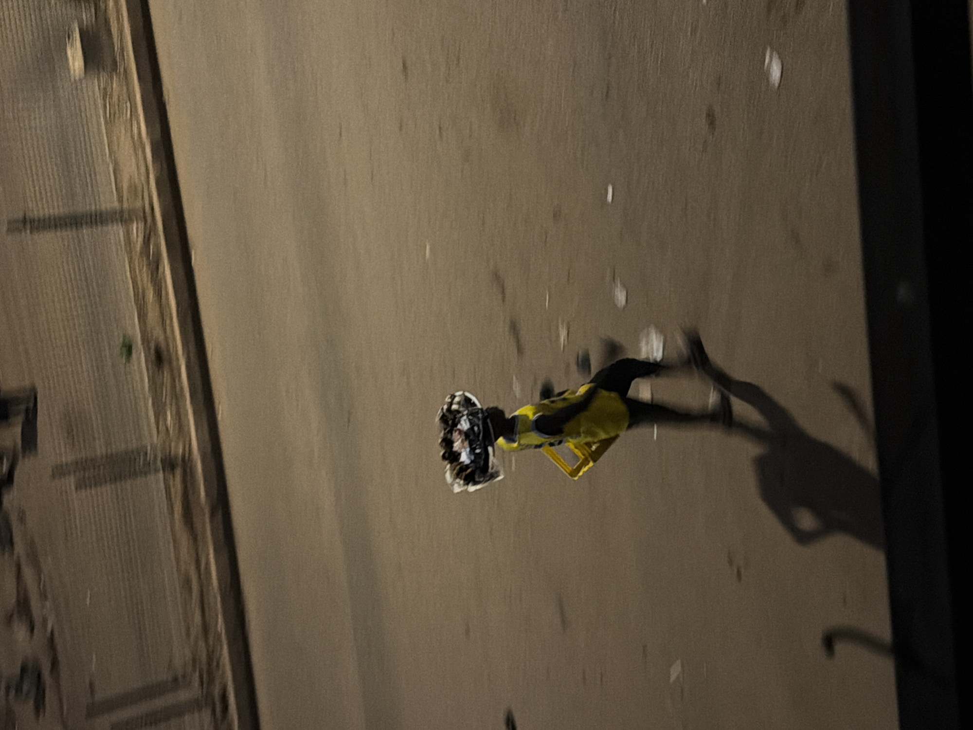 A person is walking on a paved surface, likely a road or sidewalk, in Bukuru, Nigeria. The individual, who appears to be young, is carrying a large, round basket filled with various items on their head. The basket is covered with a clear plastic material. The person is wearing a bright yellow vest over dark clothing and a yellow object, possibly a carrier or stool, is attached to their side. Their shadow is cast on the ground, indicating an overhead light source. The environment appears to be urban, with a fence and some debris visible in the background. The lighting suggests it might be evening or nighttime.