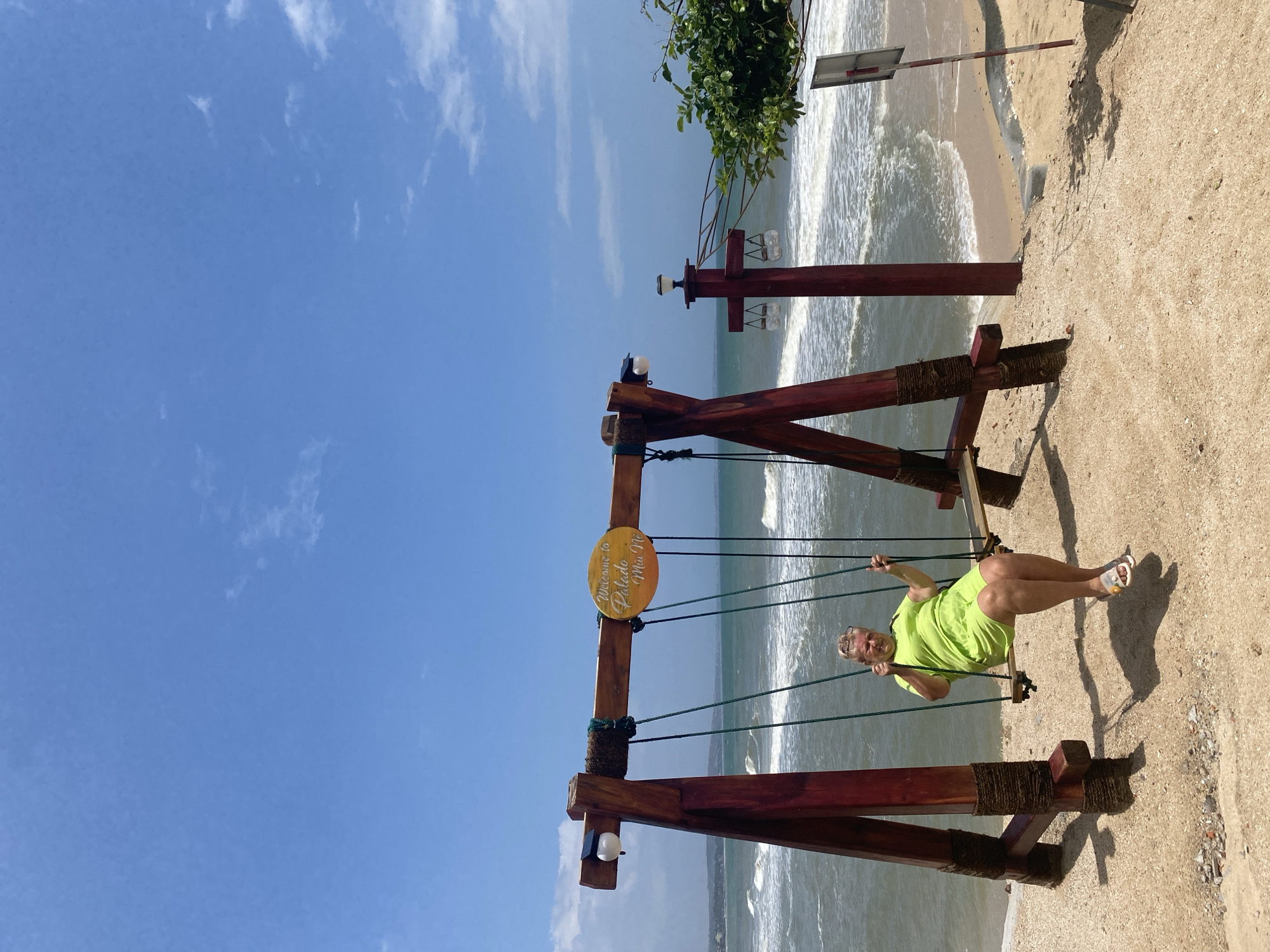 A blonde woman with graying hair, wearing a bright green outfit and white sandals, is sitting on a wooden swing at a beach in Mũi Né, Vietnam. She is holding onto the ropes of the swing with both hands and smiling at the camera. The swing set is made of dark red wooden beams and is decorated with thick rope. A yellow oval sign on the top beam of the swing reads "Welcome to Palado Mui Ne". The beach is sandy and slopes down to the ocean, where waves are breaking. The sky is blue with scattered white clouds. A tree with green leaves is visible on the right side of the image.