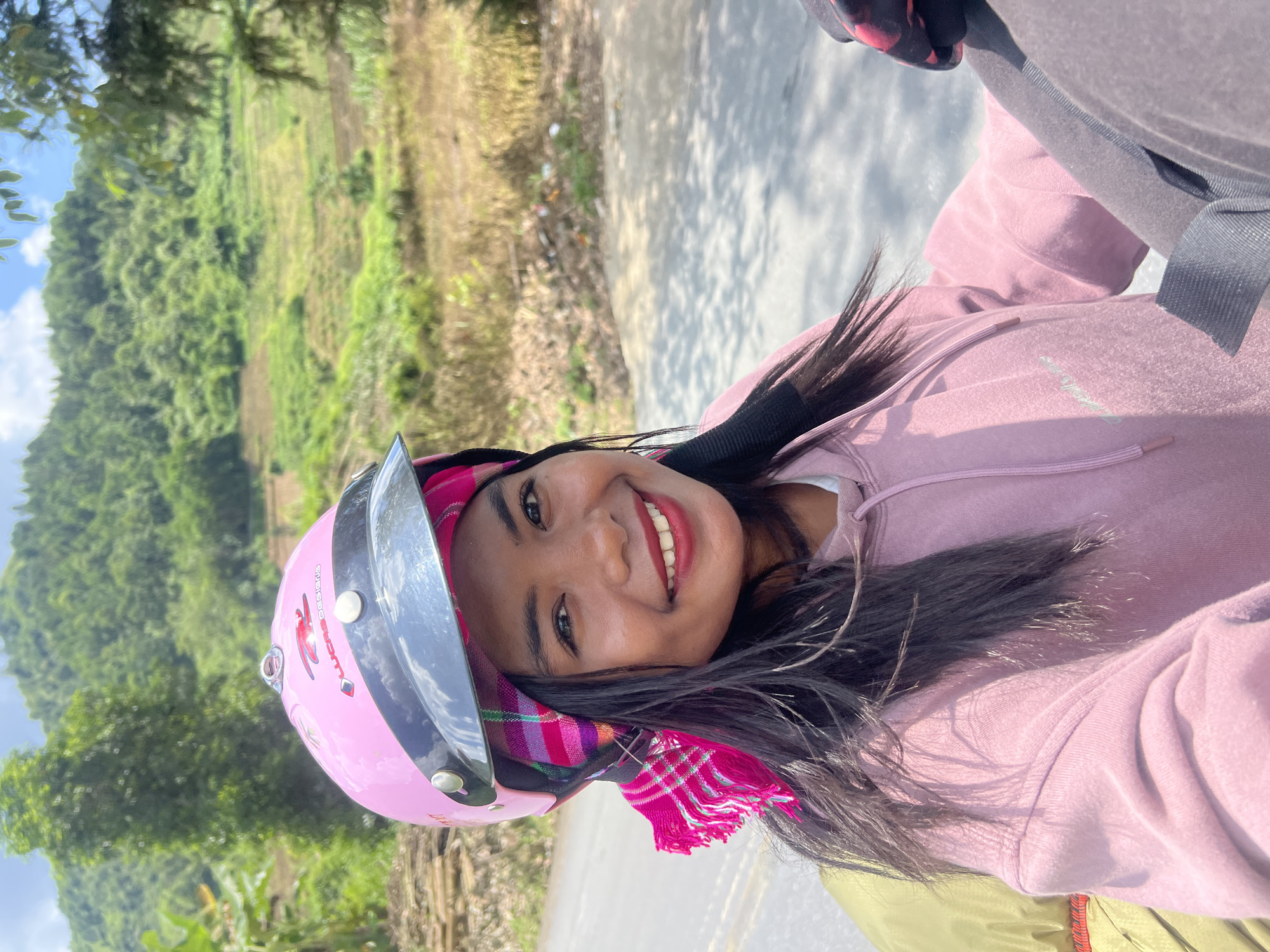 A woman wearing a pink motorcycle helmet and a pink hooded sweatshirt is captured in a close-up portrait outdoors. She has dark hair, a bright smile with red lipstick, and a pink and white checkered scarf tied around her head, with fringed ends dangling near her face. The background shows a lush, green, terraced hillside under a bright sky with some clouds, suggestive of a rural or mountainous environment in Minh Tân Commune, Vietnam. The woman appears to be seated, possibly on a motorcycle, indicated by the hint of another person's shoulder and arm in the lower right portion of the frame. The lighting suggests a sunny day.