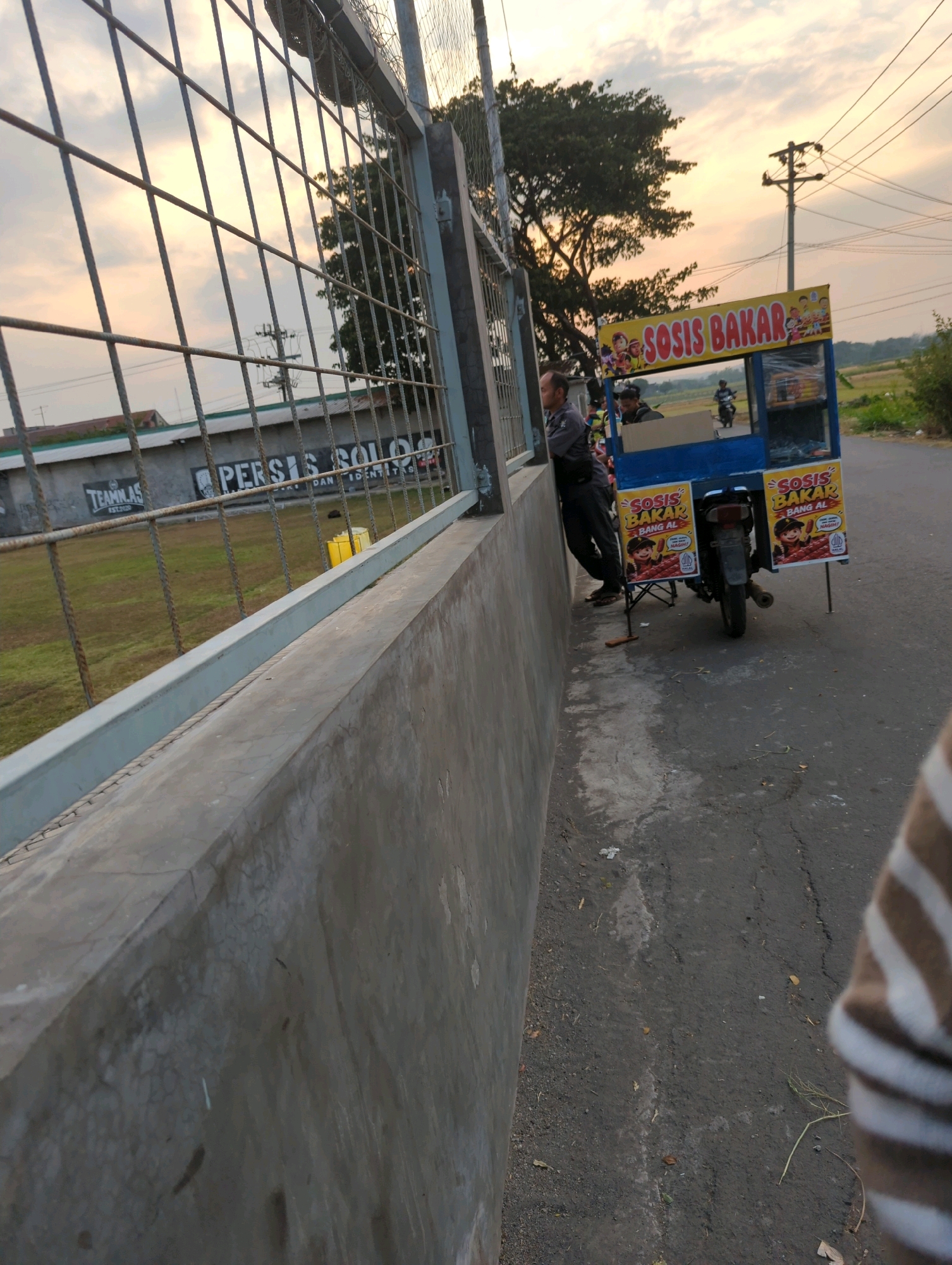 A medium shot shows a food cart selling grilled sausages on the side of a road in Andong, Indonesia. The cart, adorned with yellow and blue signage that reads "SOSIS BAKAR" (grilled sausage), is attached to a motorcycle. Two men are near the cart; one appears to be the vendor, leaning against a concrete wall, while another is operating or standing near the cart. A third person, partially visible on the right side of the frame, wears a striped shirt. The background features a grassy field behind a metal fence, a large tree, and utility poles, suggesting an outdoor setting during dusk. The overall scene captures a slice of daily life, with a focus on the mobile food business.