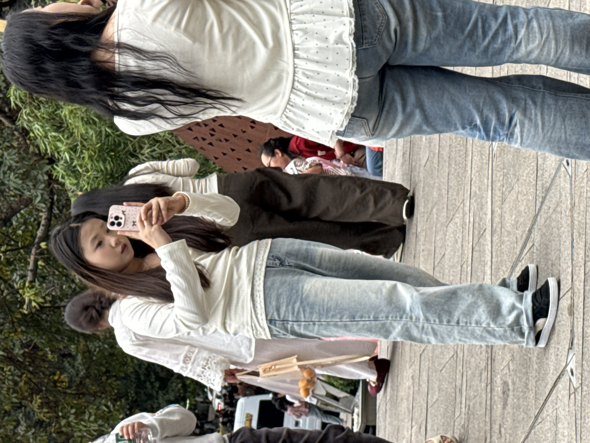 On a sunny day in Yuzhong District, China, a group of people are walking along a stone-tiled pathway outdoors. The primary focus is on a young woman with long brown hair who is holding up a smartphone to take a photograph. She is wearing a long-sleeved white shirt and light blue jeans. Behind her, another woman with long dark hair is walking away, wearing a similar white top with a ruffled hem and jeans. Other people are visible in the background, including someone holding food on a wooden stand. The environment includes green foliage to the side of the pathway, suggesting a park or garden setting.