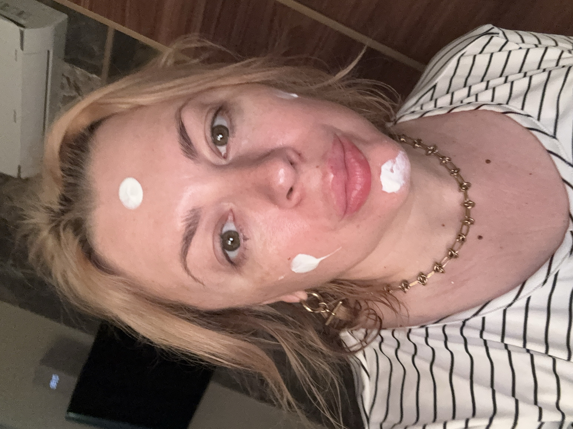 A woman with blonde hair is looking at the camera, with white cream dots on her forehead, cheeks, and chin. She is wearing a striped shirt and a gold necklace. The background appears to be an indoor setting, possibly a room, with wooden paneling and what looks like an air conditioning unit. There is no indication of the city of Sochi, Russia, in the image or its surrounding context.