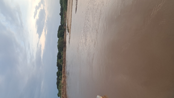 This image captures a wide, muddy-brown river in Walu, Kenya, under an overcast sky. The river occupies the majority of the frame, flowing from left to right, with its surface showing subtle current ripples. The left bank features a steep, eroded reddish-brown earthen cliff, topped with a dense line of green trees and shrubs. The right bank consists of a lower, flatter sandy-brown landmass with sparse vegetation, partially submerged. A continuous expanse of dense green trees and foliage forms the distant horizon across the background. The sky above is predominantly light gray, with a brighter, hazy area visible towards the upper right. No individuals or clearly identifiable man-made objects are present, apart from a small, indistinct light-colored object visible at the extreme left edge of the water.