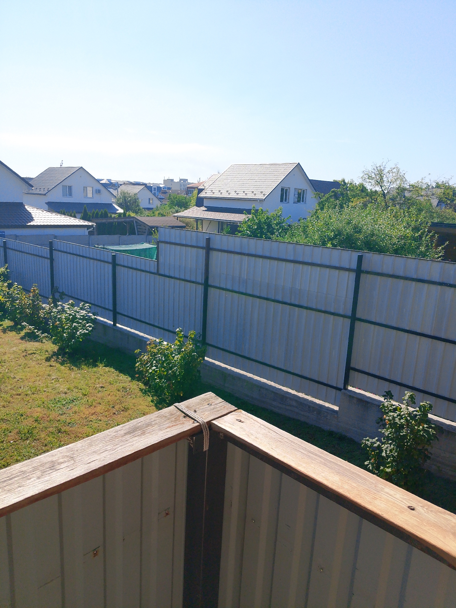 The image shows a view of a residential backyard in Irpin, Ukraine, under a clear blue sky. In the foreground, a wooden railing runs diagonally across the bottom of the frame, attached to a corrugated metal fence. Beyond the fence, several houses with tiled roofs are visible, along with trees and other vegetation. The fence itself is gray and metallic, supported by dark vertical posts. In the middle ground, a row of green bushes lines the fence. The overall scene depicts a typical suburban environment.