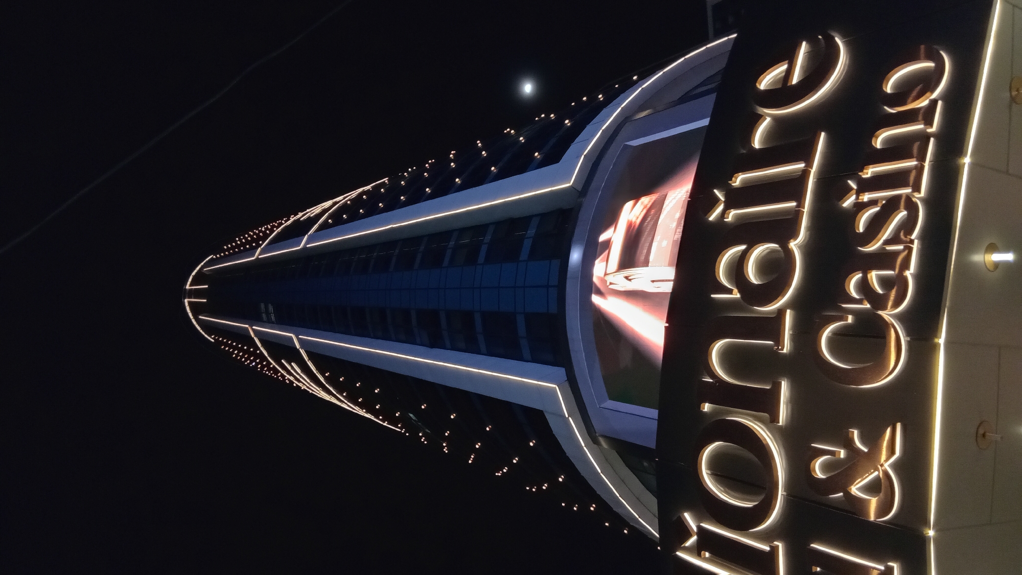 Here's a description of the image:
The image, taken at night, shows a modern building in Batumi, Georgia. The "Colosseum Marina Hotel" is depicted with its illuminated signage. The building's design features a cylindrical shape with decorative lights along its edges. Part of the building's glass facade is visible. The sky is dark, with what appears to be the moon visible.