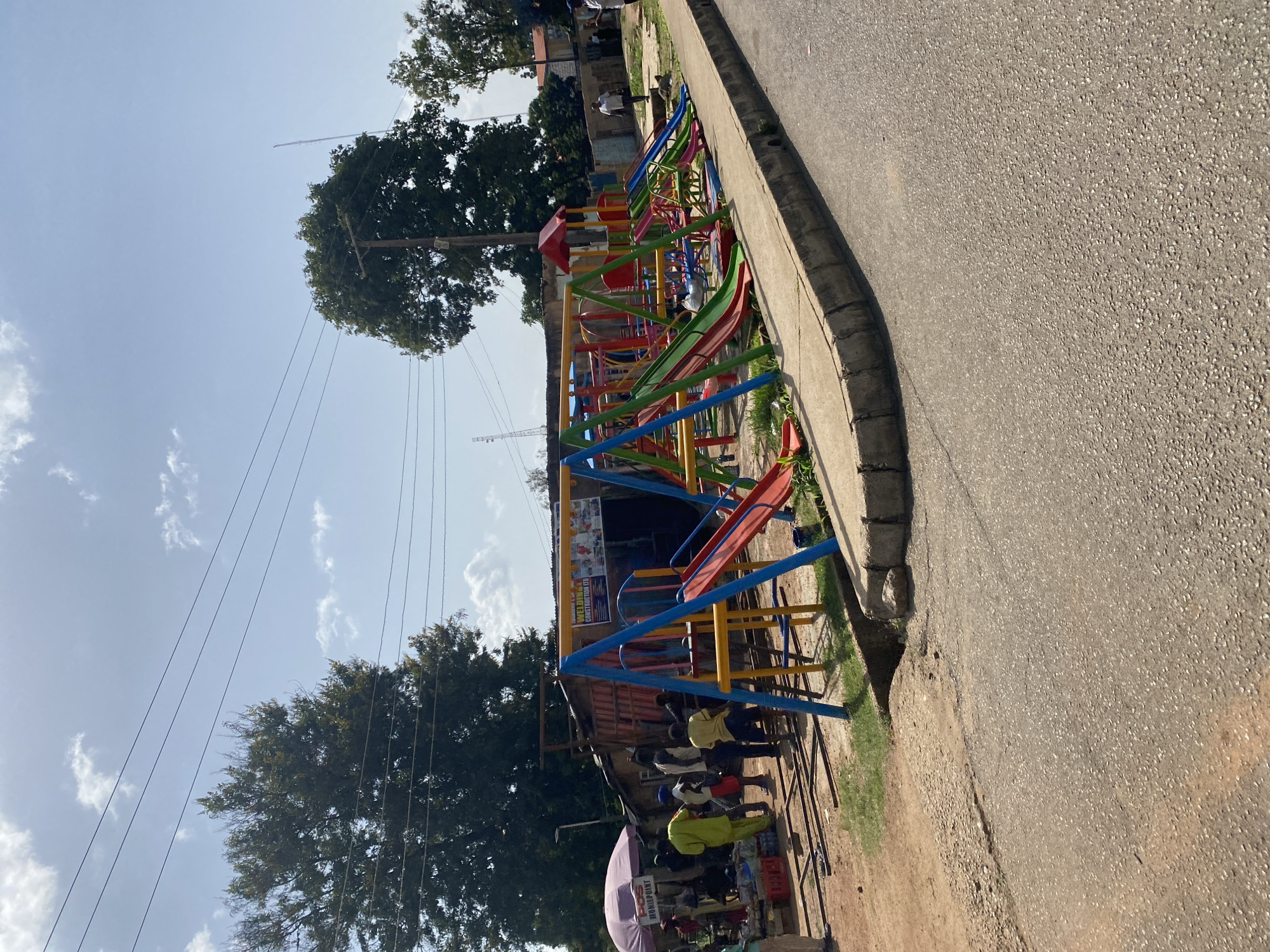 The image depicts an outdoor scene in Jos, Nigeria, featuring a brightly colored playground with swings and slides situated alongside a dirt road and a sidewalk. To the left of the playground, a small market stall with a purple umbrella displays various goods, and several people are gathered around it, appearing to be engaged in commerce. Behind the market stall, a makeshift structure, possibly a shop or dwelling, is visible, constructed from corrugated metal and wood. Further in the background, trees, buildings, and utility poles with overhead wires suggest an urban or semi-urban environment. The ground is a mix of asphalt, dirt, and grass, with a concrete curb running along the edge of the road. The sky is partly cloudy, indicating a daytime scene.
