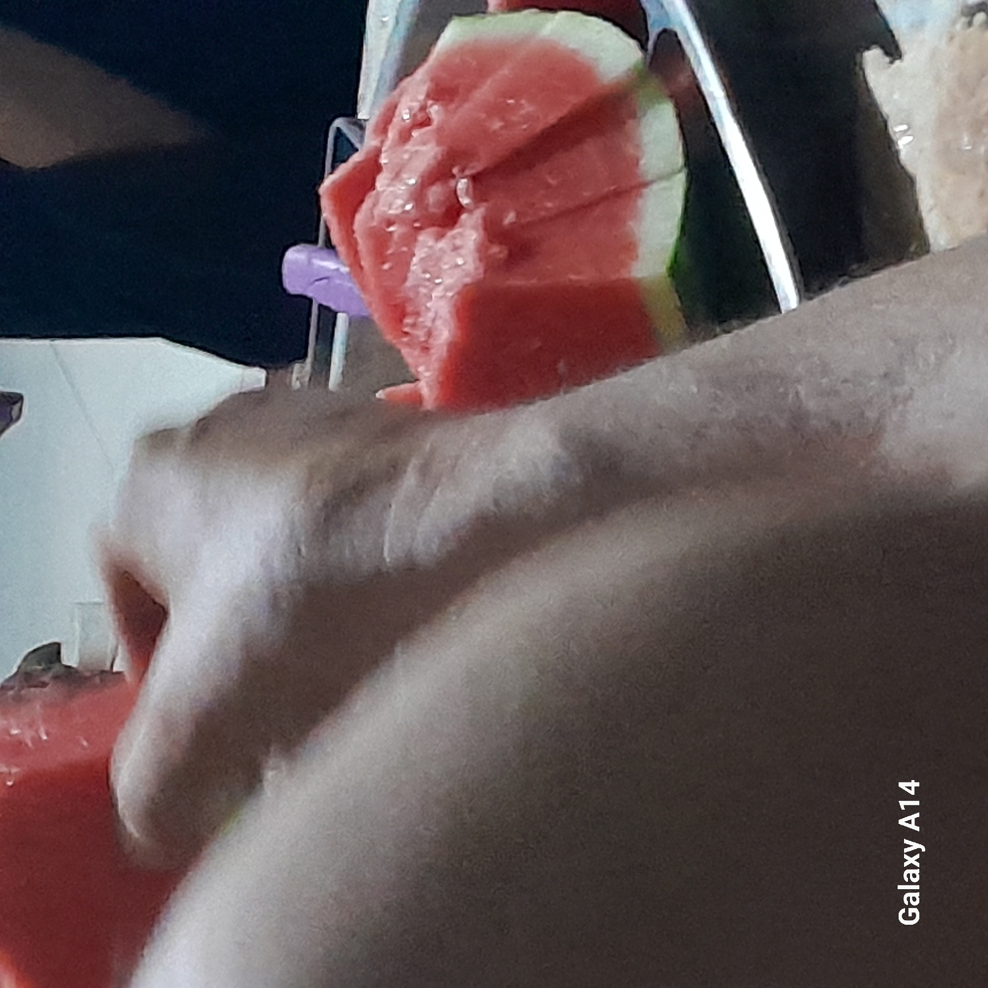 An extreme close-up shot shows a person's arm and a plate of sliced watermelon. The arm, in the foreground, is light-skinned and shows some texture and a few faint marks. The watermelon slices are a vibrant red and white, with some juice visible on the surface, arranged in a circular metal bowl. A purple utensil is partially visible near the watermelon. The background is blurred, with dark clothing and a white surface suggesting an indoor setting. The text "Galaxy A14" is present in the lower left corner, indicating the device used to capture the image. The overall scene is candid and focuses on the act of eating watermelon. The location of Khan Yunis, Palestinian Territories, is not visually discernible from the image itself.