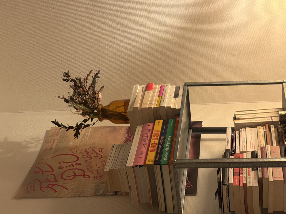 The image depicts a static interior scene within a corner of a room, identified as being located in Paris, France. A grey, industrial-style metal shelving unit is prominently featured, with its top shelf occupying the center-left. This shelf is laden with numerous stacks of books, including titles such as "Yves Saint Laurent," "WARHOL - BASQUIAT," "Bacon en toutes lettres," "Jakob + MacFarlane," and "DONATION PERRON & ARTISTES," primarily in French. To the right of these books, an amber-colored glass bottle, functioning as a vase, holds an arrangement of dried botanical branches and foliage. Behind the vase and the upper book stacks, leaned against the light-colored, textured walls, is a large, light brown poster. This poster features prominent red-pink script, including Japanese characters translating to "The day I died" and partially visible English text "Virgin Suicide?". The scene is illuminated by warm, artificial light originating from the right, casting soft shadows. The lower sections of the shelving unit are also filled with densely packed books. No individuals or actions are present in the frame.
