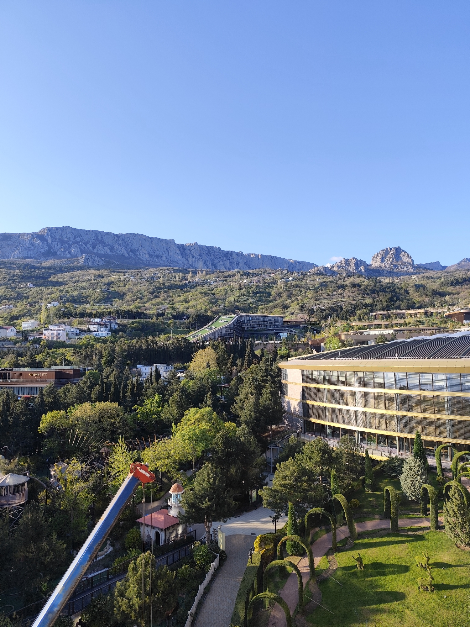 This image presents a panoramic view of a scenic mountainous landscape interspersed with modern architecture and meticulously maintained gardens, all under a clear blue sky.
In the background, a majestic range of rugged, rocky mountains stretches across the horizon, with portions covered in verdant vegetation. The middle ground is characterized by rolling green hills densely covered with various types of trees, including many conifers, and dotted with numerous residential houses.
Several contemporary buildings are prominent in the foreground and mid-ground. To the right, a large building with a distinctive golden-colored facade featuring multiple glass windows and horizontal louvers stands out. On the left side, another modern structure is visible, displaying the text "MHIYA LIFE" on its exterior. Further up the hillside, a building with a unique green, sloping roof can be seen.
The lower part of the image reveals beautifully manicured gardens. These include lush green lawns, winding pathways, and elaborate topiary art, such as trimmed archways and stylized animal figures. A bright blue and orange arm of a construction crane extends diagonally from the bottom left corner into the scene, indicating nearby construction or maintenance activities. Smaller decorative structures, including a gazebo and a charming lighthouse-like building, are nestled amongst the trees.
The scene is illuminated by bright sunlight, suggesting either early morning or late afternoon, with long shadows cast by the trees and buildings. The sky is a pristine, cloudless blue. The overall atmosphere is peaceful and picturesque, indicative of a resort or upscale residential area seamlessly integrated into a stunning natural environment. The visual cues align with the provided location context of Симеизский поселковый совет, Ukraine. No individuals are visible in the image.