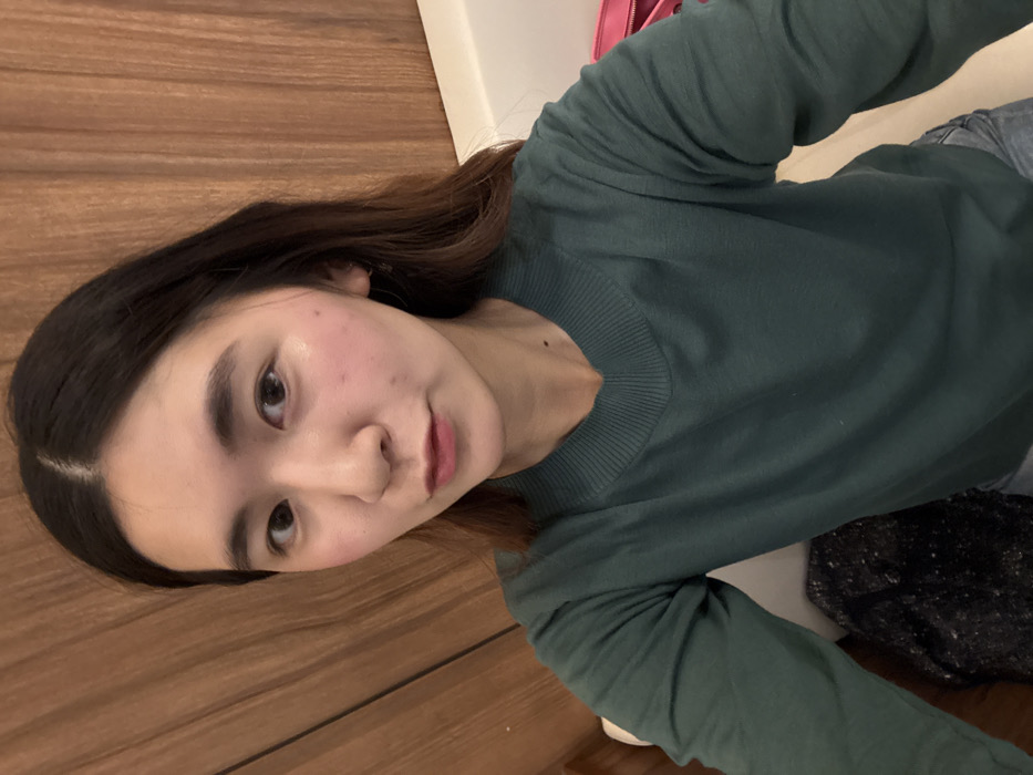 A young East Asian female is depicted in a close-up, chest-up shot. She has long, dark brown hair parted centrally and wears a dark teal, long-sleeved V-neck sweater. Her eyes are open, and she looks directly at the camera with a neutral expression. Her cheeks exhibit a subtle flush. The background features a vertically paneled wooden wall. To the subject's right, a white surface, possibly an armrest, is visible, alongside a partial view of a pink object. A dark, textured item occupies the lower-left foreground. The indoor setting lacks discernible landmarks or unique architectural features to identify a specific location in Paris, France.