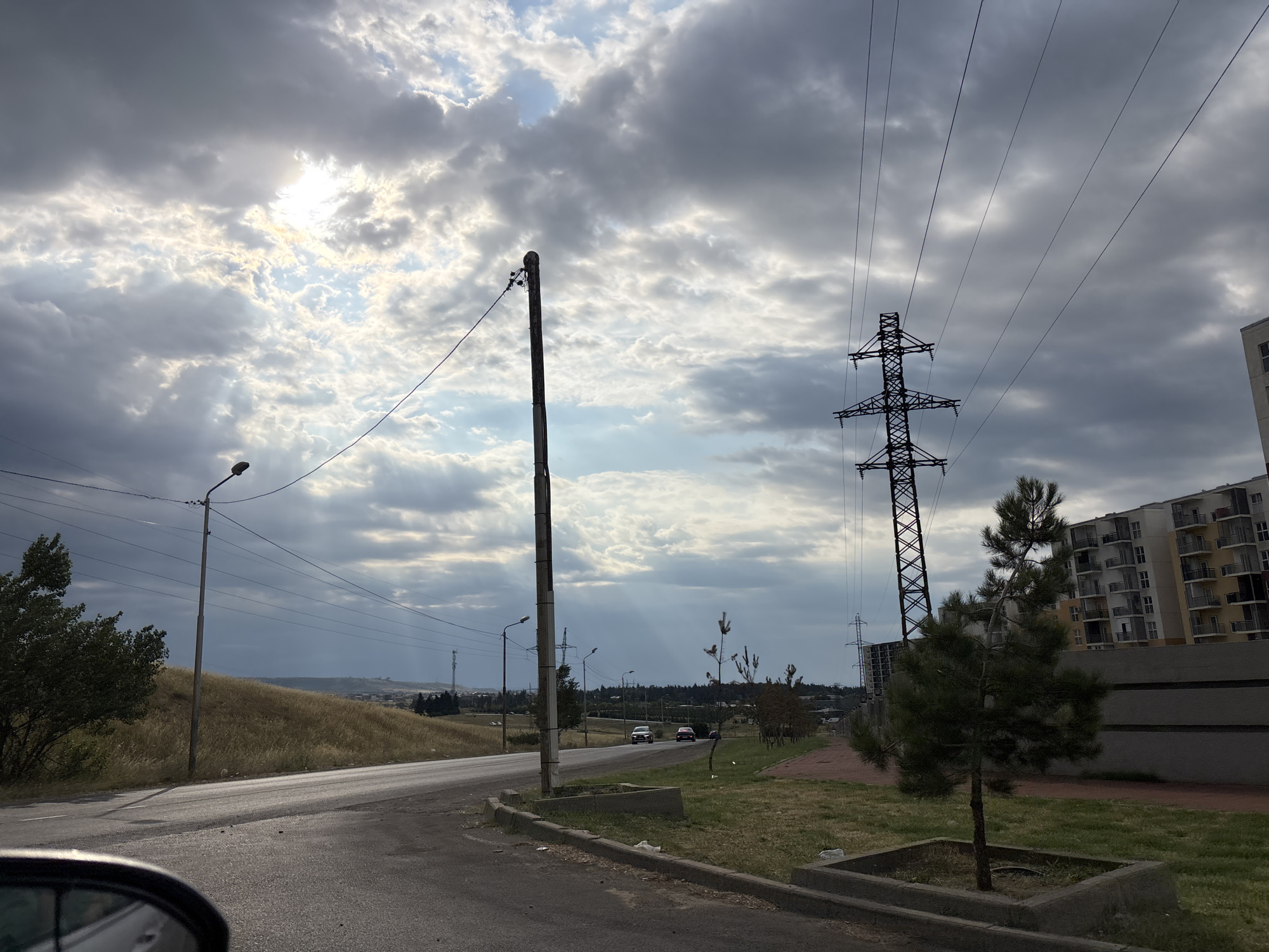 The image shows a road with a view of a residential building and a power line tower in Tbilisi, Georgia. The sky is overcast with some sunlight peeking through the clouds. The road is paved and curves to the left. There is a grassy area with a small tree and a concrete planter to the right of the road. A multistory building with balconies is visible in the background. A large power line tower with several wires extends across the sky. A utility pole with a street light is also present near the road. The scene appears to be an urban or semi-urban environment.