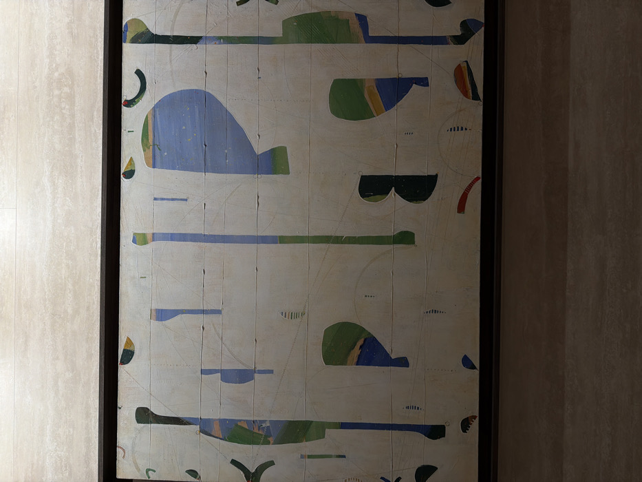 A large, rectangular abstract painting is centered on a light beige, horizontally-banded stone wall in New York City, United States of America. The artwork, enclosed in a dark, flat frame, features a predominantly off-white or light beige background divided into several horizontal panels. It displays a variety of irregular, geometric, and organic shapes in shades of blue, green, yellow, orange, and occasional red and black. Prominent forms include elongated vertical elements, rounded 'P'-like and 'B'-like shapes, and crescent fragments. Subtle dotted or stitched lines traverse sections of the canvas, adding texture. The wall surface exhibits natural stone striations. The scene is brightly and evenly lit. No individuals, actions, or direct interactions are depicted.