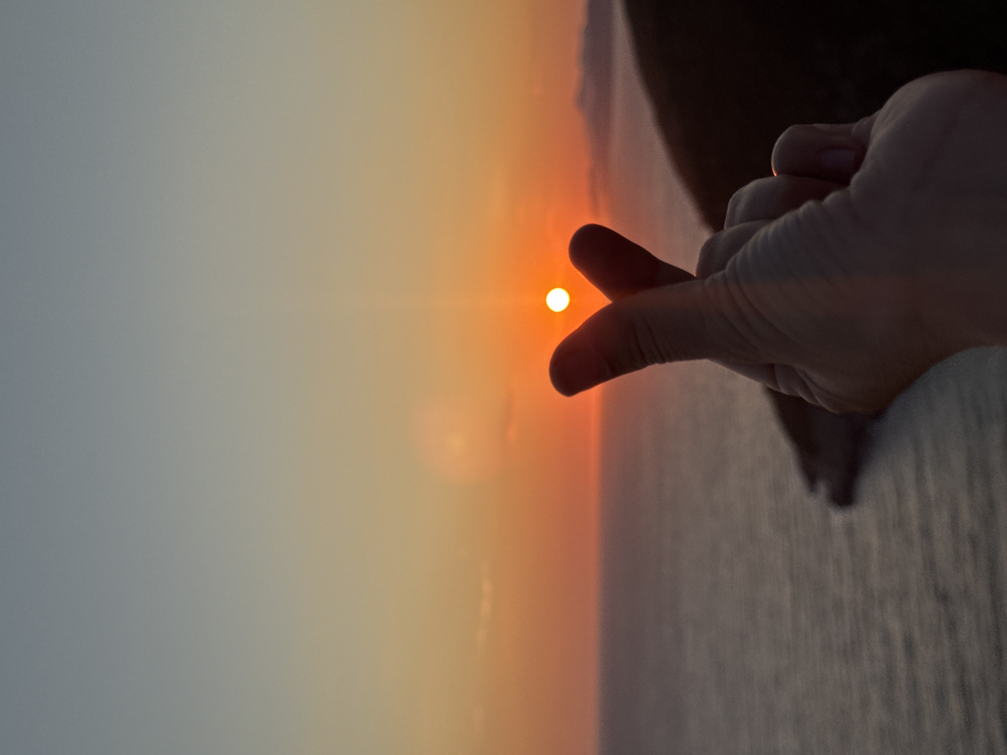 The image shows a hand forming a heart shape in front of a sunset over the Adriatic Sea. The sun is a bright orb low on the horizon, casting an orange glow across the sky and water. The hand is in the foreground, with the thumb and index finger creating the heart shape. In the background, distant mountains are visible. The scene is set in Zagora, Montenegro, a coastal town known for its beautiful sunsets. The overall mood is romantic and peaceful.