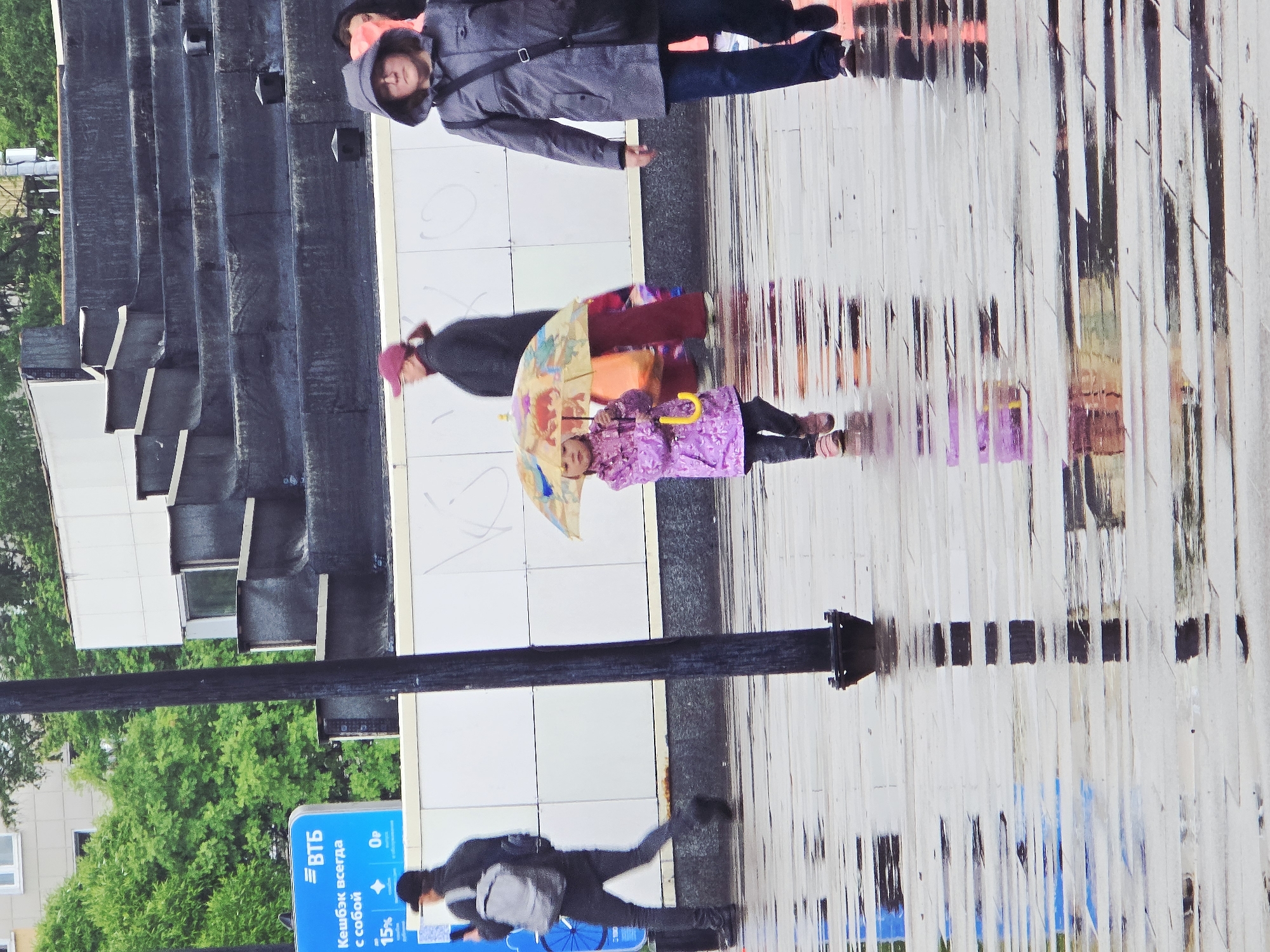 The image depicts a rainy scene on a sidewalk in Moscow, Russia. A woman in a grey coat and blue jeans walks parallel to a younger girl in a purple outfit holding a colorful umbrella. A person wearing a dark jacket and a pink hat is positioned to the left of the child. The sidewalk is wet, with reflective patches mimicking people and objects. A pedestrian with a light grey backpack walks in the lower quadrant, toward the bank, where a bank advertisement is posted in the bottom left corner of the image. A dark pole is next to the sign. A building with alternating light and dark panels appears on the left side of the photo, and greenery is visible behind the building.
