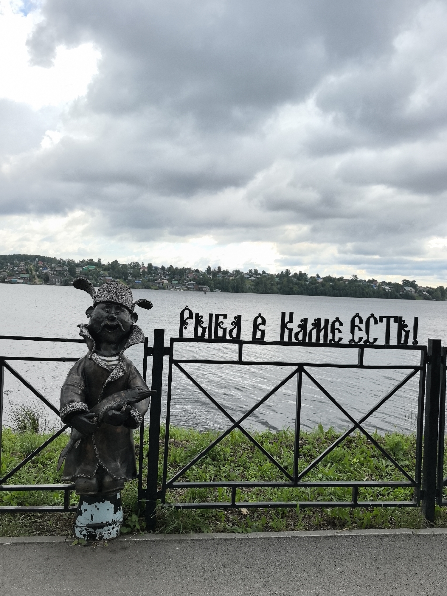 The image depicts a bronze sculpture of a whimsical character standing on a pedestal in front of a body of water. The sculpture is of a short, stout figure wearing a hat with ear-like flaps and a coat with fur trim, holding a fish. To the right of the sculpture, a black metal fence with decorative elements runs along the water's edge. On the fence, the Cyrillic text "РЫБА В КАМЕ ЕСТЬ!" is visible, which translates to "There is fish in Kamen!". In the background, across the water, is a shoreline with houses and trees, indicating a settlement. The sky is overcast with gray clouds. The scene is likely a public waterfront area in the city of Dobryanka, Russia, a place known for its fishing traditions.
