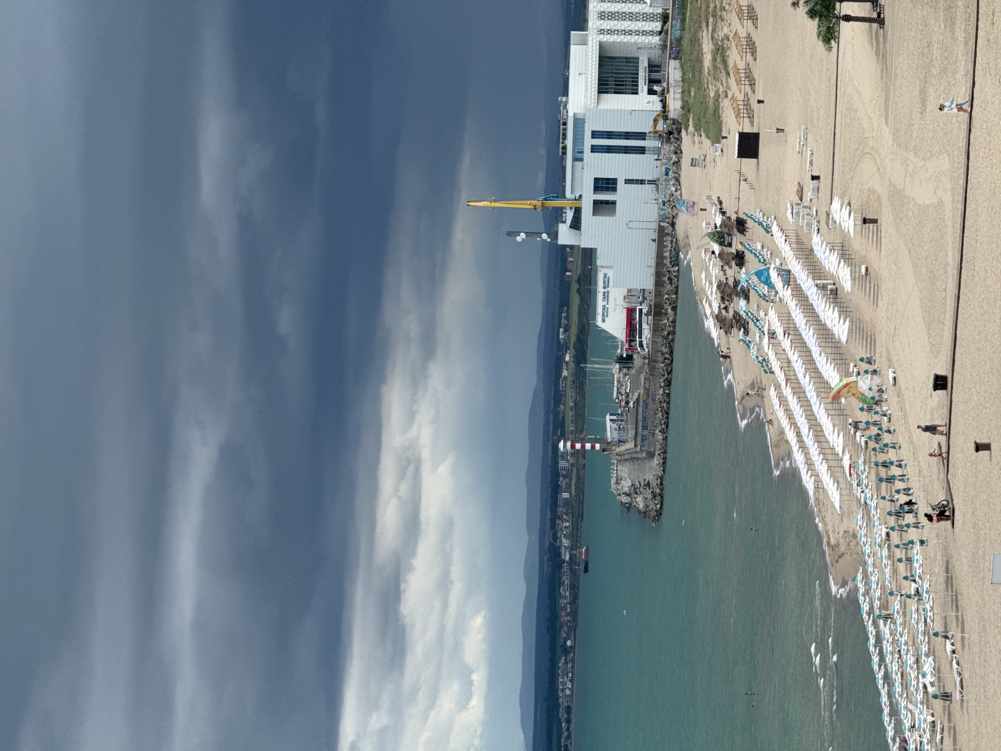 The image depicts a view of the Burgas Sea Garden and its beach in Burgas, Bulgaria. The scene is characterized by a dark, overcast sky, suggesting impending weather. The foreground is dominated by a sandy beach with numerous rows of white and blue beach umbrellas, many of which are open, indicating a moderately populated beach area. Several beach chairs are visible beneath the umbrellas.
Along the waterline, the turquoise sea shows gentle waves and a few scattered individuals can be seen swimming or wading. The beach extends towards a man-made pier. On the pier, a red and white striped lighthouse stands prominently, adjacent to a modern, white building complex with glass facades and a large yellow crane extending upwards. This building appears to be a terminal or port facility, with the text "МОРСКА ГАРА БУРГАС" (Sea Station Burgas) visible on its side.
Further out in the sea, a small red vessel is visible. The background shows a hazy coastline with hills and more distant structures, suggesting the urban sprawl of Burgas. The overall impression is a coastal area preparing for or experiencing inclement weather, with a mix of recreational beachgoers and industrial port infrastructure.