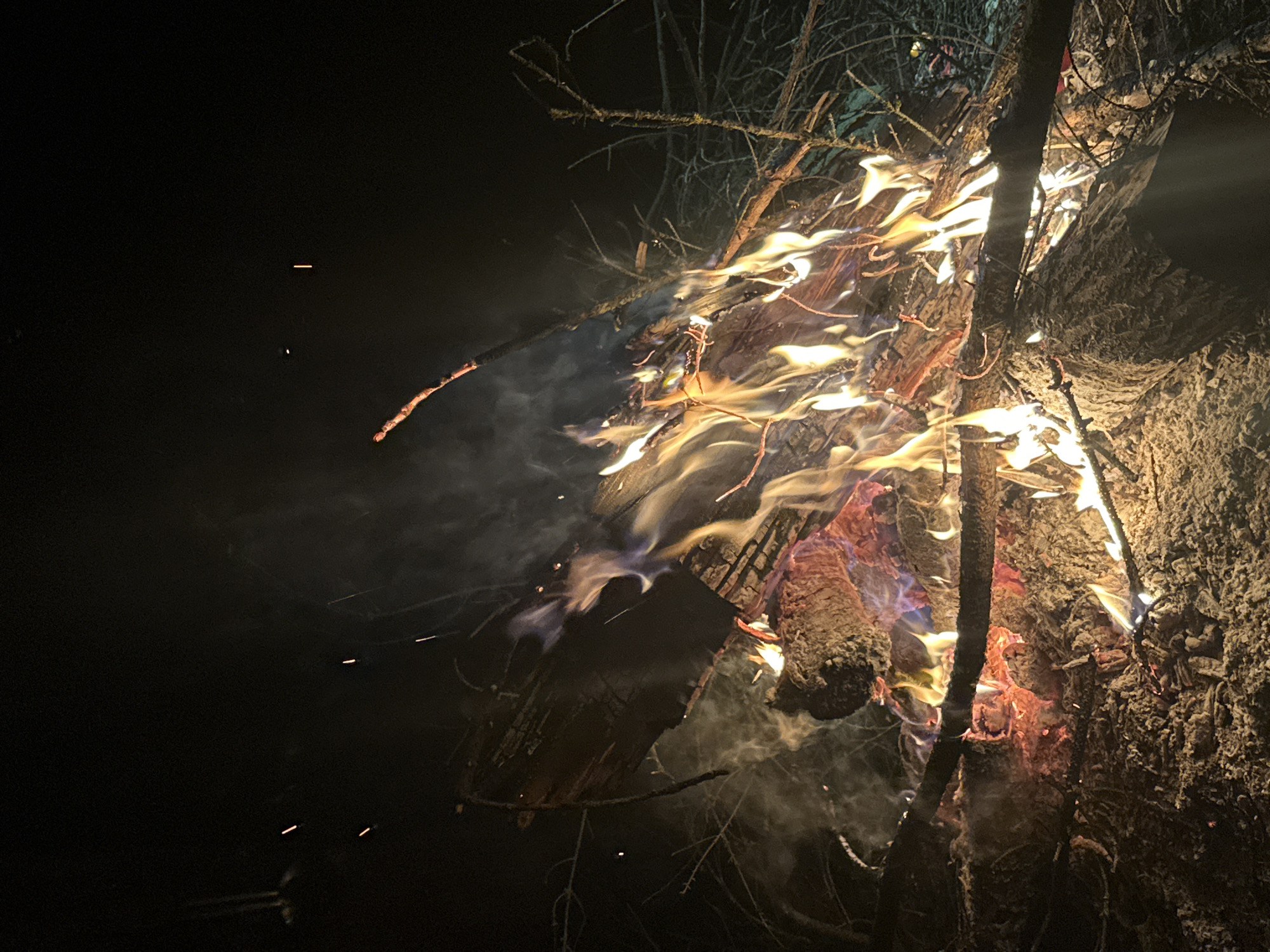 The image depicts a close-up view of a roaring bonfire at night. The fire is contained within what appears to be a stack of logs and branches. Flames rise and swirl, displaying various colors from yellow and orange to hints of blue. Smoke is visible, particularly towards the left side of the frame. Sparks emanate from the fire. The surrounding environment is dark, but textures of the logs and tree branches can be discerned. The specific location within Юзуфоўскі сельскі Савет, Belarus, is not discernible from the image.