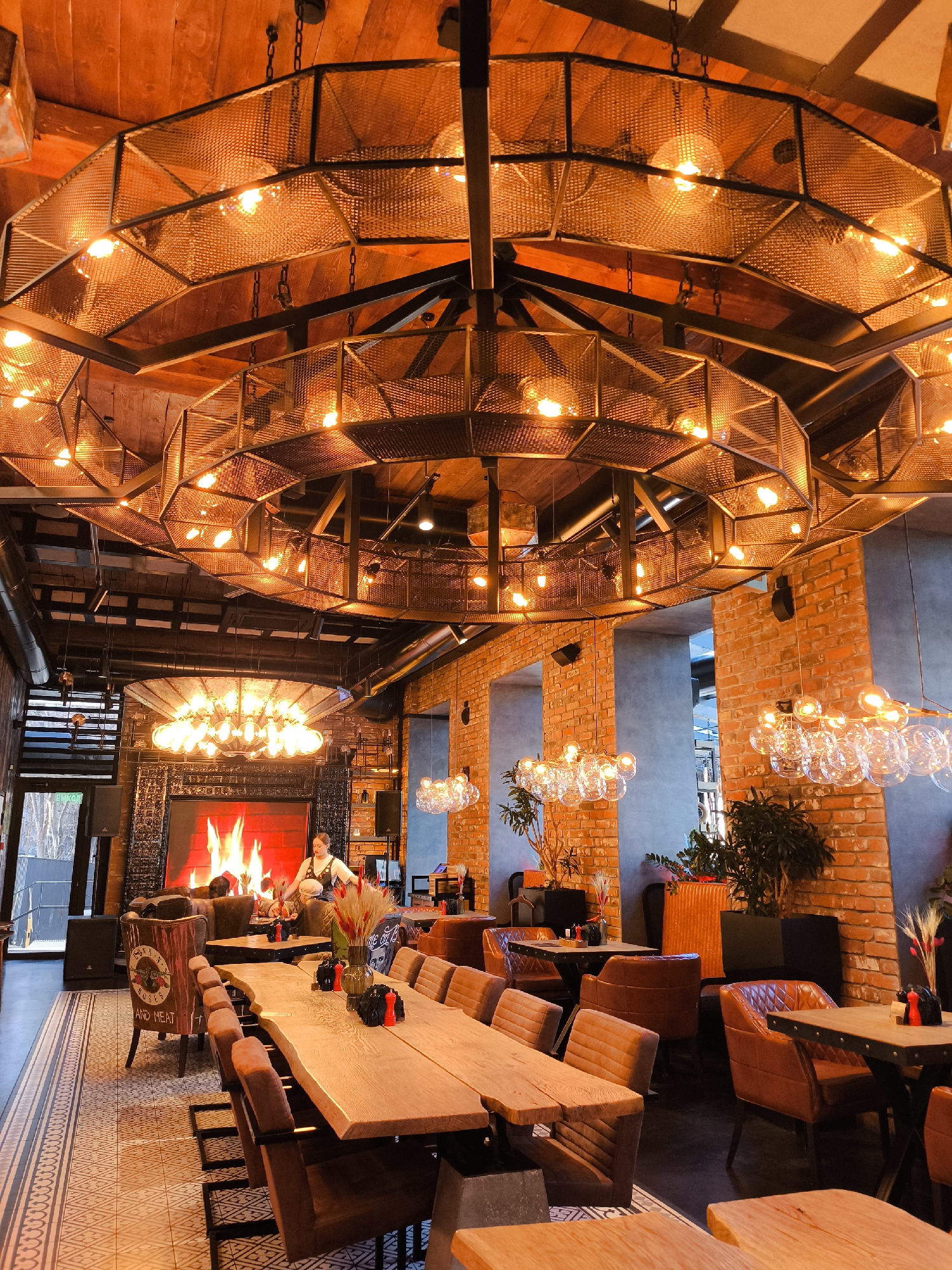 The image displays the interior of the "Guns N Roses Bar & Meat" restaurant, located at Svobody Street, 173, Izhevsk, Russia. The setting features a rustic industrial aesthetic, characterized by a wooden plank ceiling, exposed red brick walls, and sections of dark gray concrete. The floor consists of a patterned geometric tile pathway transitioning to darker, solid flooring beneath dining areas.
Overhead, multiple large, dark-metal mesh chandeliers with numerous exposed filament bulbs provide warm, amber illumination. Additional smaller chandeliers with clear spherical glass bulbs hang lower. In the background, a prominent rectangular digital fireplace screen displays realistic flames within a dark, textured surround.
A long, live-edge wooden communal table, set with dark pepper and salt grinders and a vase of dried red and beige plumes, occupies the midground. It is flanked by multiple dark brown upholstered chairs with metal frames; some chair backs display the "GUNS N ROSES AND MEAT" logo. Various smaller square tables with matching dark brown armchairs are distributed throughout the space.
A single female individual with light-colored hair, dressed in a dark top with light straps, stands near the digital fireplace, possibly tending to a table or observing the display. Potted green plants are strategically placed throughout the restaurant in dark and orange planters. Large black audio speakers are visible near the fireplace and a glass doorway on the far left, which offers a glimpse of a winter exterior with bare trees. The restaurant appears largely unoccupied by customers.