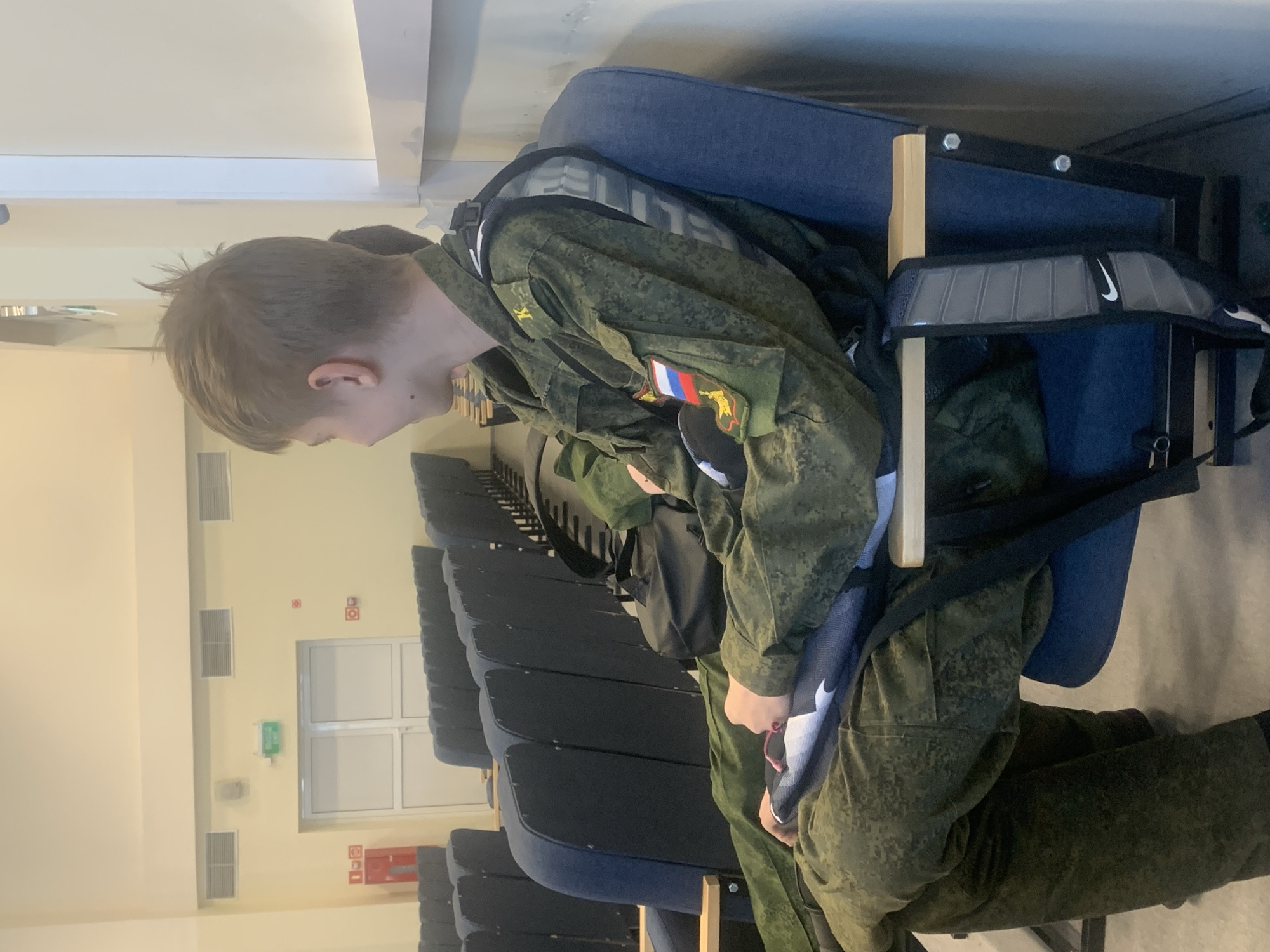 A young person in military fatigues is seated in a dark blue auditorium chair. They have a backpack draped on the chair behind them, with a visible Nike logo. The person is facing left, looking down at their hands. A Russian flag patch is visible on the uniform. The setting is an auditorium with rows of empty dark blue chairs. The background shows a wall with a door and some safety equipment, including a fire hose reel and a green emergency exit sign. The image was taken in Chaikovsky, Russia.