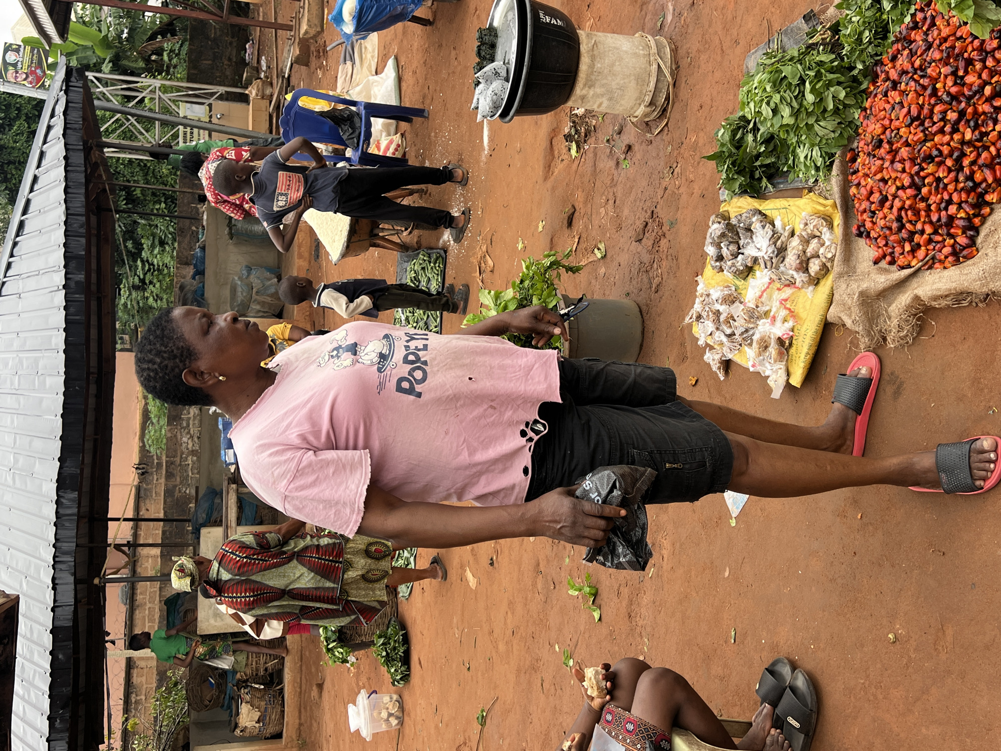 An outdoor market scene is depicted in Nkpor, Nigeria, with a focus on a woman standing center frame. She is wearing a pink t-shirt with a "Popeye" graphic and black shorts, and pink-and-black slide sandals. Her right hand clutches a small black plastic bag. To her left, a person is partially visible, seated and wearing a patterned skirt, holding a piece of food. In the foreground, a pile of red palm nuts is spread on a burlap sack, and to their right, a large bunch of green leafy vegetables is arranged on a dark fabric. Various other items, including more vegetables, baskets, and bags of produce, are scattered throughout the marketplace. Several children are also present, one wearing a blue t-shirt with a flag design, and another in a green shirt. The ground is a reddish-brown dirt. Overhead, a metal-roofed structure provides shade. The overall impression is a busy, everyday market environment.