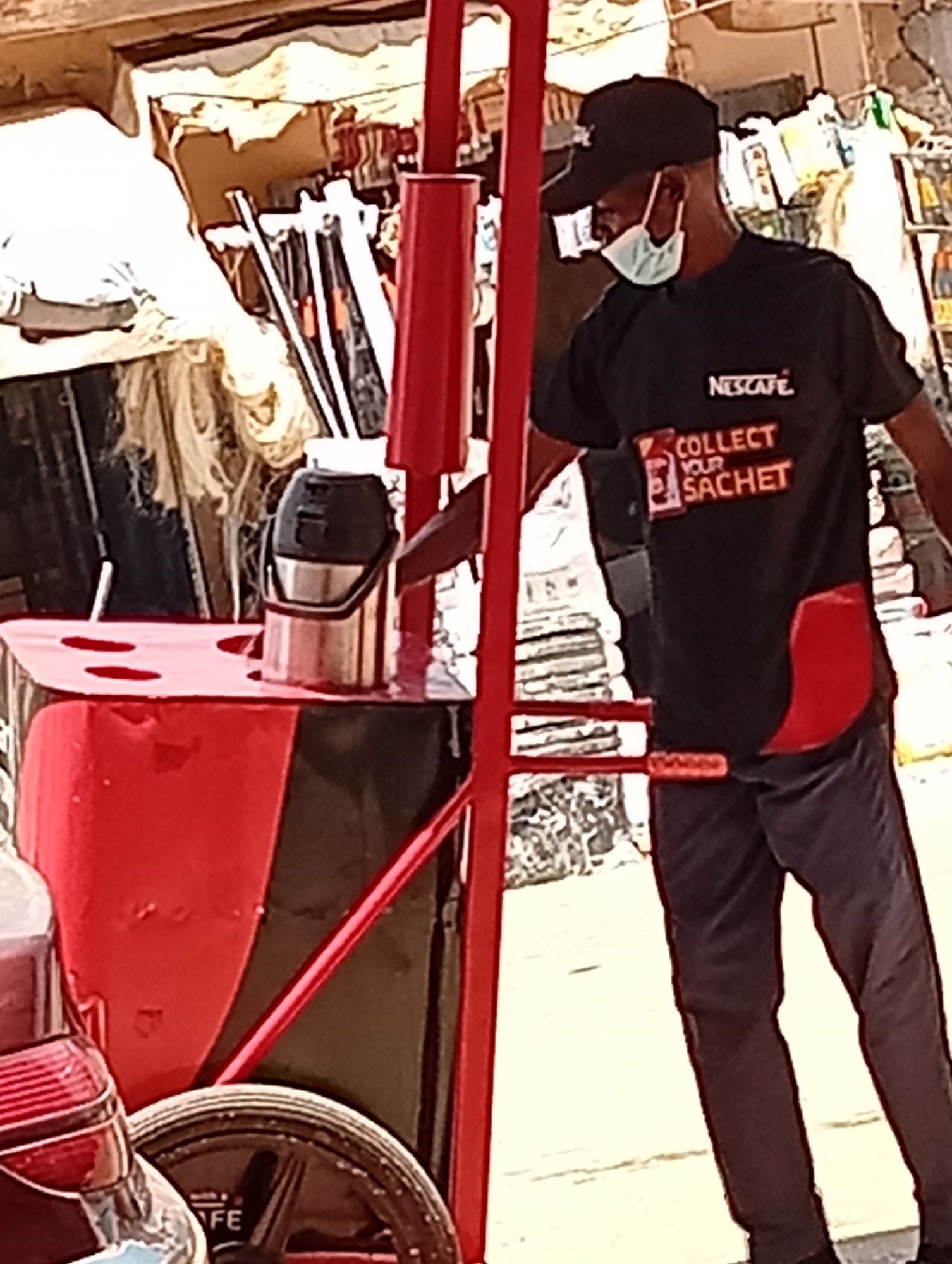 In Maiburiji, Nigeria, a vendor wearing a black "NESCAFE" t-shirt, dark pants, a baseball cap, and a face mask stands next to a red cart. The cart includes a thermos, several straws, and a wheel with the letters "FE" visible. The vendor is positioned on a city street. Background elements include merchandise displayed in an outdoor retail setting.