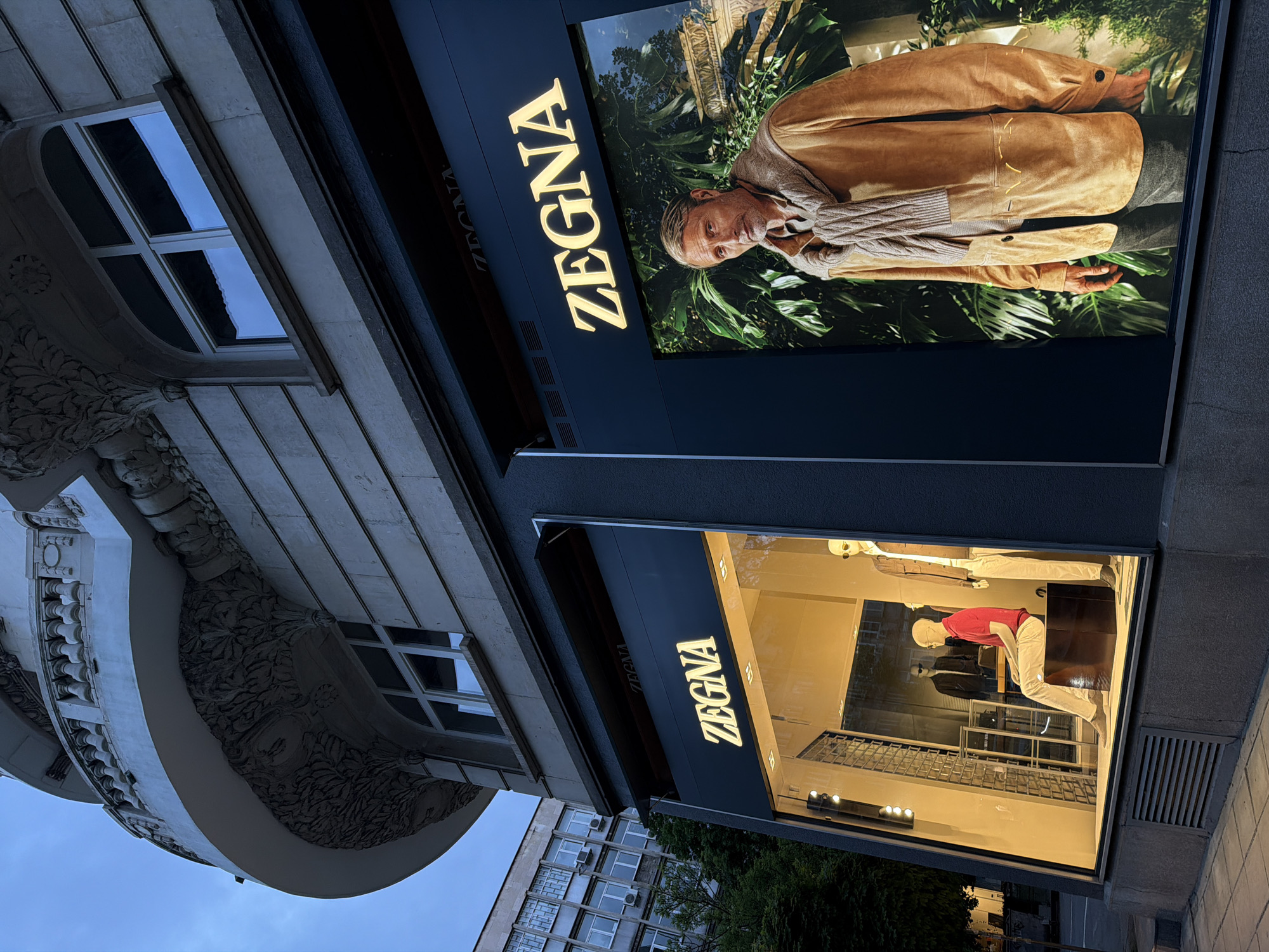 The image captures a ZEGNA store, likely in Sofia, Bulgaria, given the architectural style of the adjacent building. The storefront features two illuminated ZEGNA signs, with a large advertisement above the window display. The advertisement depicts a man in a brown jacket and scarf against a backdrop of green foliage. The window display showcases mannequins dressed in ZEGNA clothing, including one in a red shirt and light-colored trousers. The surrounding buildings display a mix of architectural styles, including older ornamented structures and modern apartment buildings. The sky is visible in the background, suggesting early evening or late afternoon light.