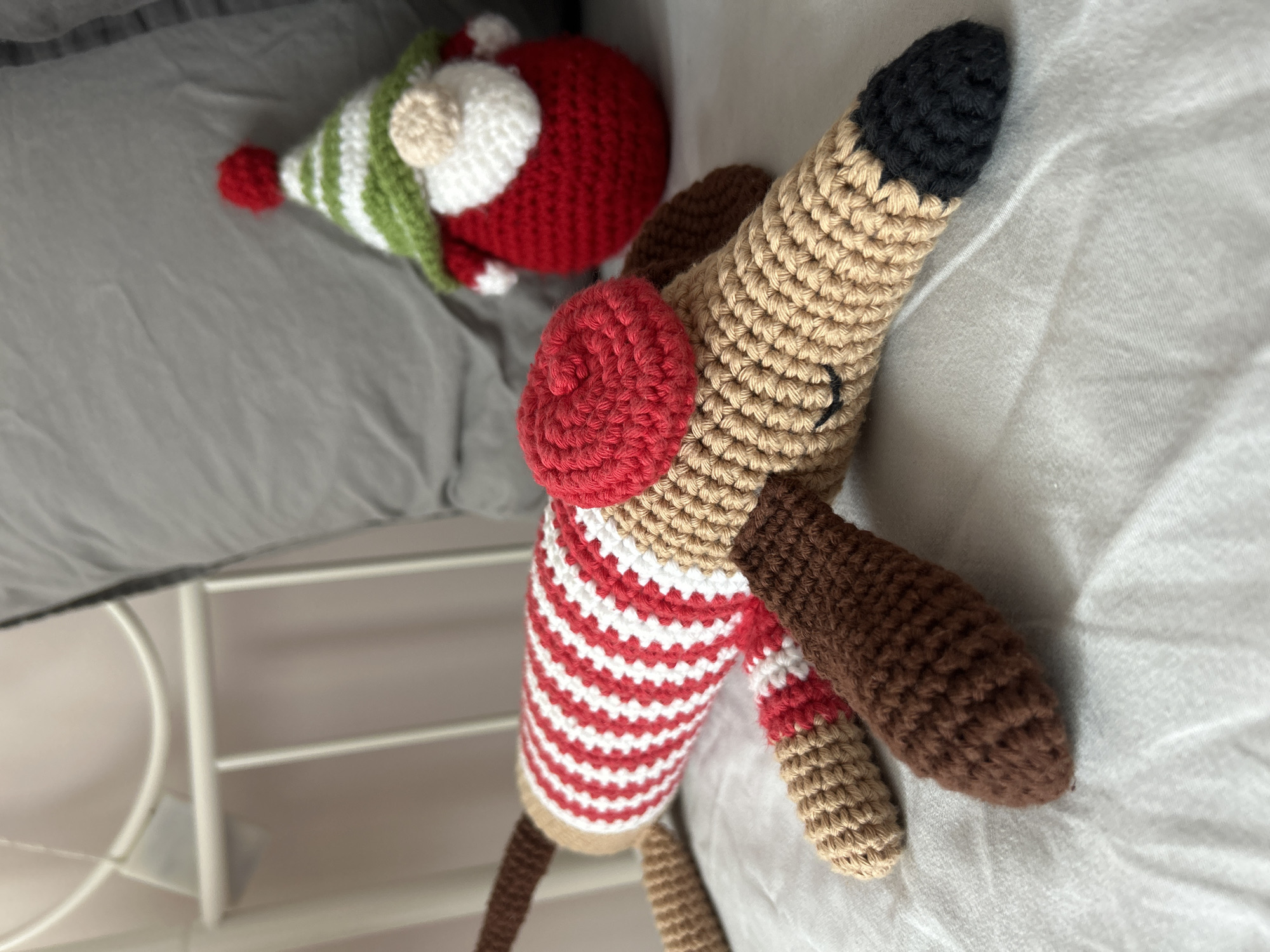 The image shows two crochet toys, a dog and a gnome, resting on a bed covered with gray and white sheets. The dog toy has a tan body, brown ears and legs, a black nose, and a red and white striped shirt. The gnome toy has a red and white body, green and white hat, and a beige pom-pom. The location is likely indoors, possibly in a home or hotel, in Plovdiv, Bulgaria.