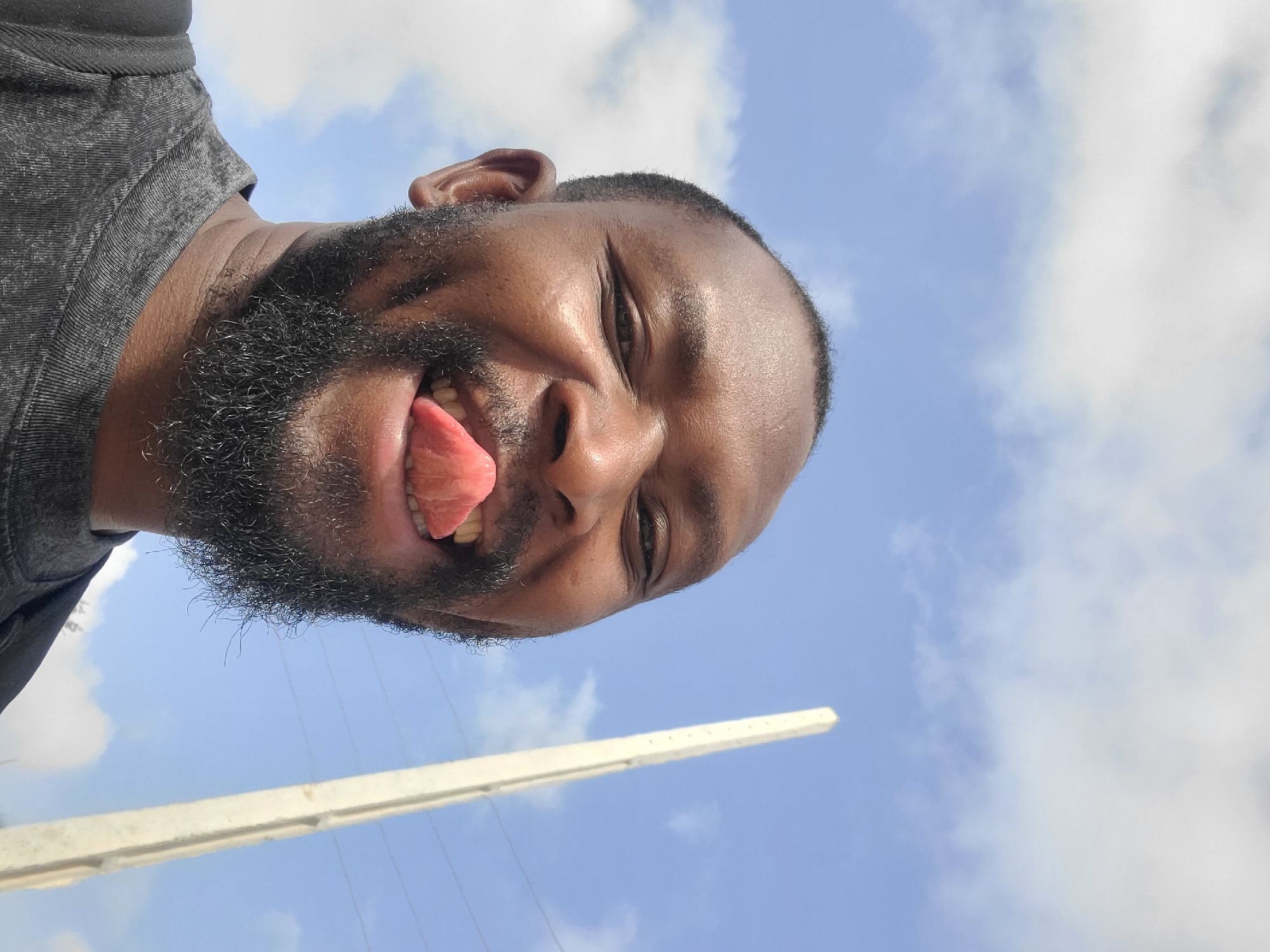 A man with dark skin, a full beard, and short dark hair is photographed from a low angle against a backdrop of a blue and cloudy sky. He is sticking his tongue out and smiling. The man is wearing a gray shirt. In the lower part of the frame, a white, concrete utility pole is visible. The location is determined to be Barakin Akawo, Nigeria.