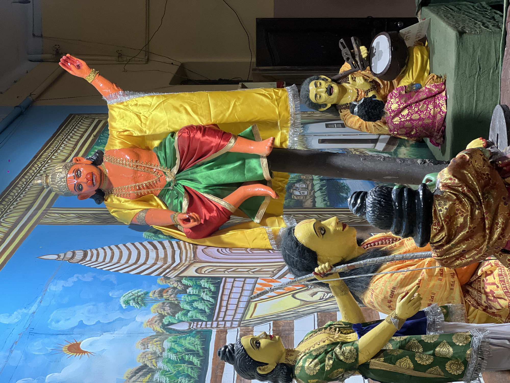 The image depicts a tableau of painted figures, likely part of a religious display, possibly in Varanasi, India. Hanuman, painted orange, lies on a yellow fabric, gesturing with his right arm. He wears a green and red lower garment. Behind him, a painted backdrop depicts a temple structure against a blue sky with trees. To the right, two female figures painted in yellow and wearing traditional garments hold a bow and arrow, suggesting a narrative scene. Behind them, a man in yellow with a black beard and carrying a drum is seated. The tableau appears to be indoors, with elements of a room, including a television, visible in the background. The overall scene is colorful and suggests a devotional or artistic setting.