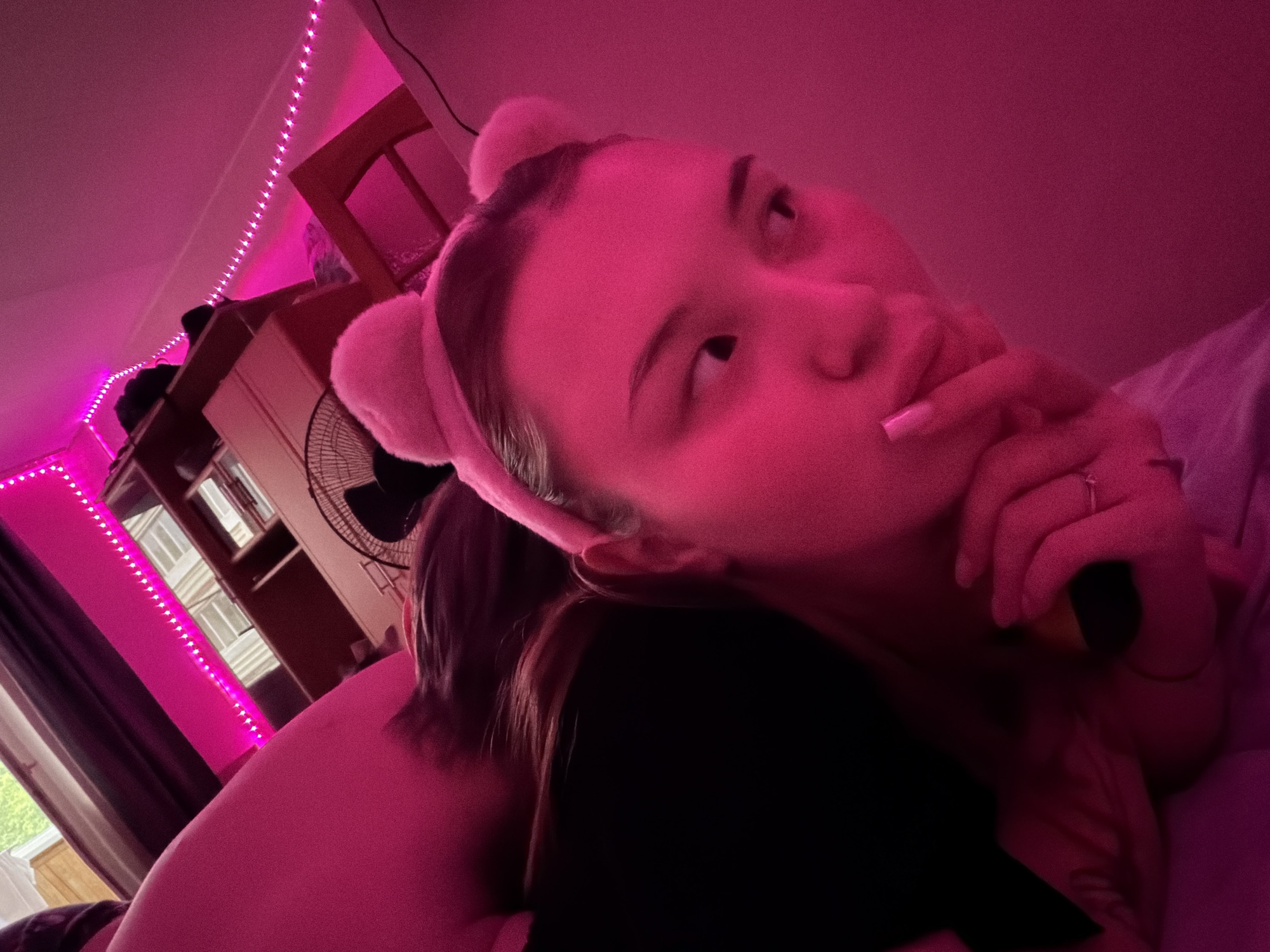 A young woman with a pink bear-ear headband is lying down, looking up and to her left, with a finger to her lips. She is wearing a ring on her finger. Part of a person's shoulder and black shirt is visible next to her. Behind them is a bookshelf, a fan, and a wooden chair. Pink string lights are visible on the wall. The location is unknown.