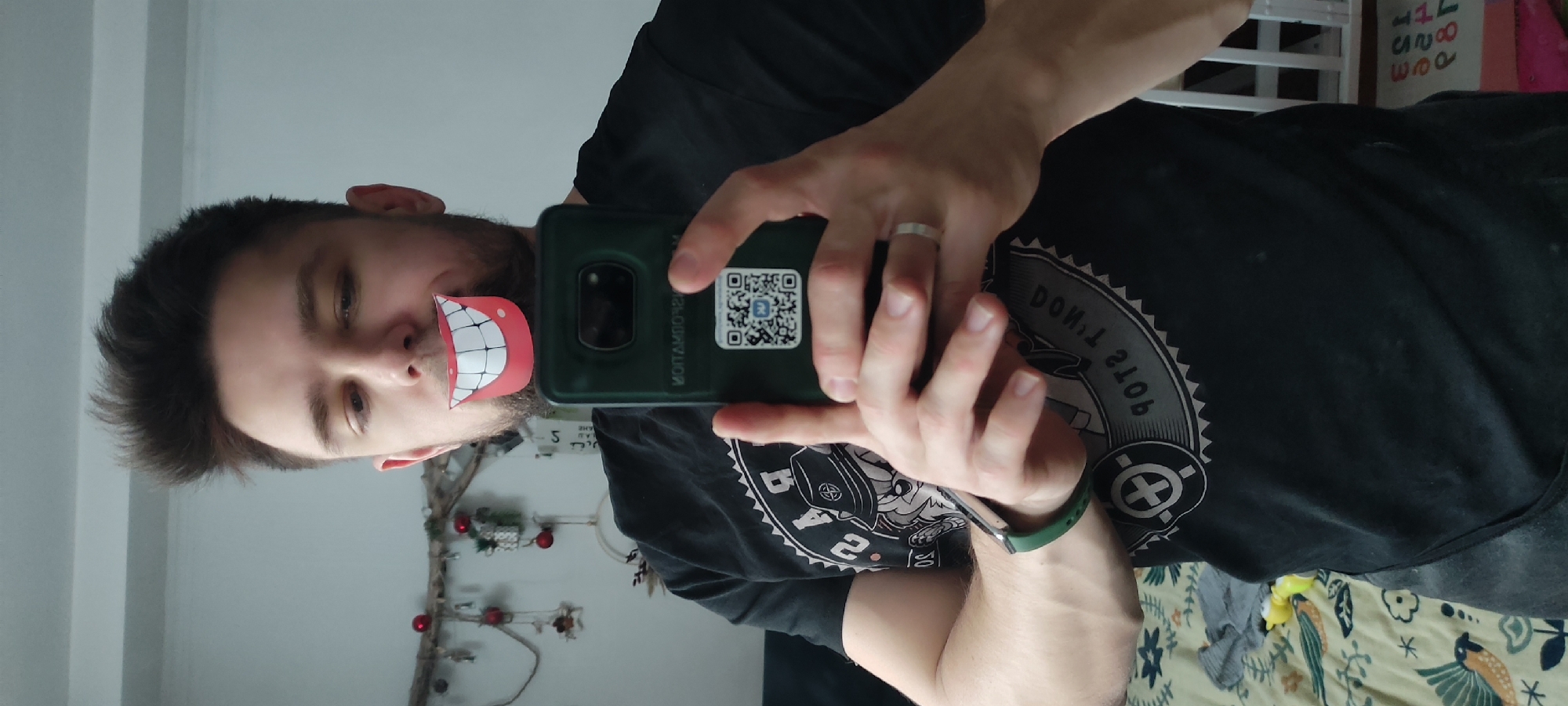 A male is taking a selfie with a phone that has a green case with a QR code on it. The male is wearing a black t-shirt with a graphic design and a wedding band on his left ring finger. He has a paper cutout of a mouth with white teeth taped over his mouth. The location is likely in an interior in Fryazino, Russia. A Christmas decoration is in the background, along with what appears to be a radiator.