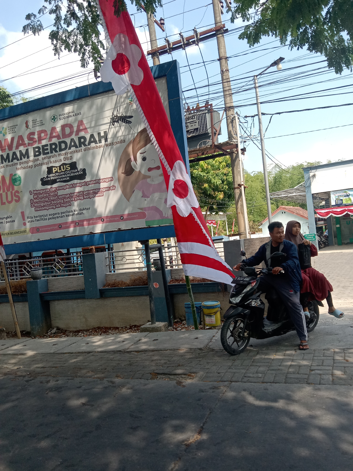 The image shows a street scene in Sugihwaras, Indonesia, with a large billboard on the left, a utility pole with electrical wires and a transformer in the background, and a red and white striped flag hanging from a pole in the foreground. A man and a woman are riding a black motorcycle in the middle ground, heading towards the right side of the image. The man is driving, and the woman is a passenger. The billboard has text in Indonesian, including "WASPADA MAM BERDARAH" which translates to "BEWARE OF BLOODY MAM," and an illustration of a mosquito and a distressed child. The environment appears to be a public area, possibly near a health clinic given the billboard's content about health warnings. The weather is clear and sunny.