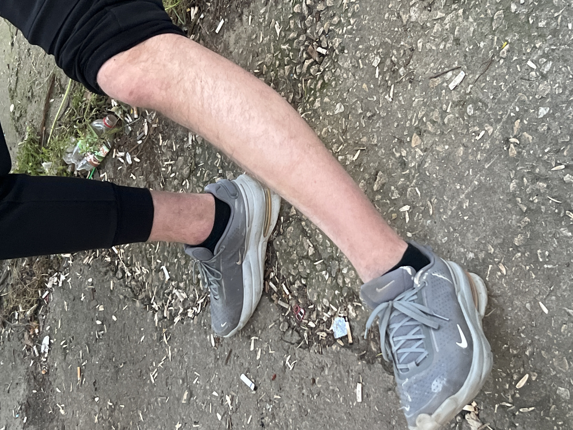 The image captures a ground-level view of the lower legs and feet of two individuals on a littered concrete or asphalt surface in the city of Dobrich, Bulgaria. The ground is covered with numerous white and yellowed cigarette butts, a substantial quantity of light-colored, elongated husks consistent with sunflower seed shells, and various small pieces of debris, including a red metallic pull-tab and fragments of foil. In the upper right quadrant, two clear plastic bottles are visible among sparse, dry vegetation.
One individual, positioned on the left side of the frame, is standing. They are wearing black athletic trousers and a grey Nike sneaker with a white sole and black socks. The second individual, positioned on the right, is in a crouched or kneeling posture, with their right leg extended forward and bent at the knee. This individual also wears black athletic trousers, revealing a bare, hairy lower leg. The visible knee exhibits a reddish discoloration. Their right foot, clad in a grey Nike sneaker with a white sole and black socks, rests on the ground towards the lower left of the image. No direct interaction between the two individuals is depicted.