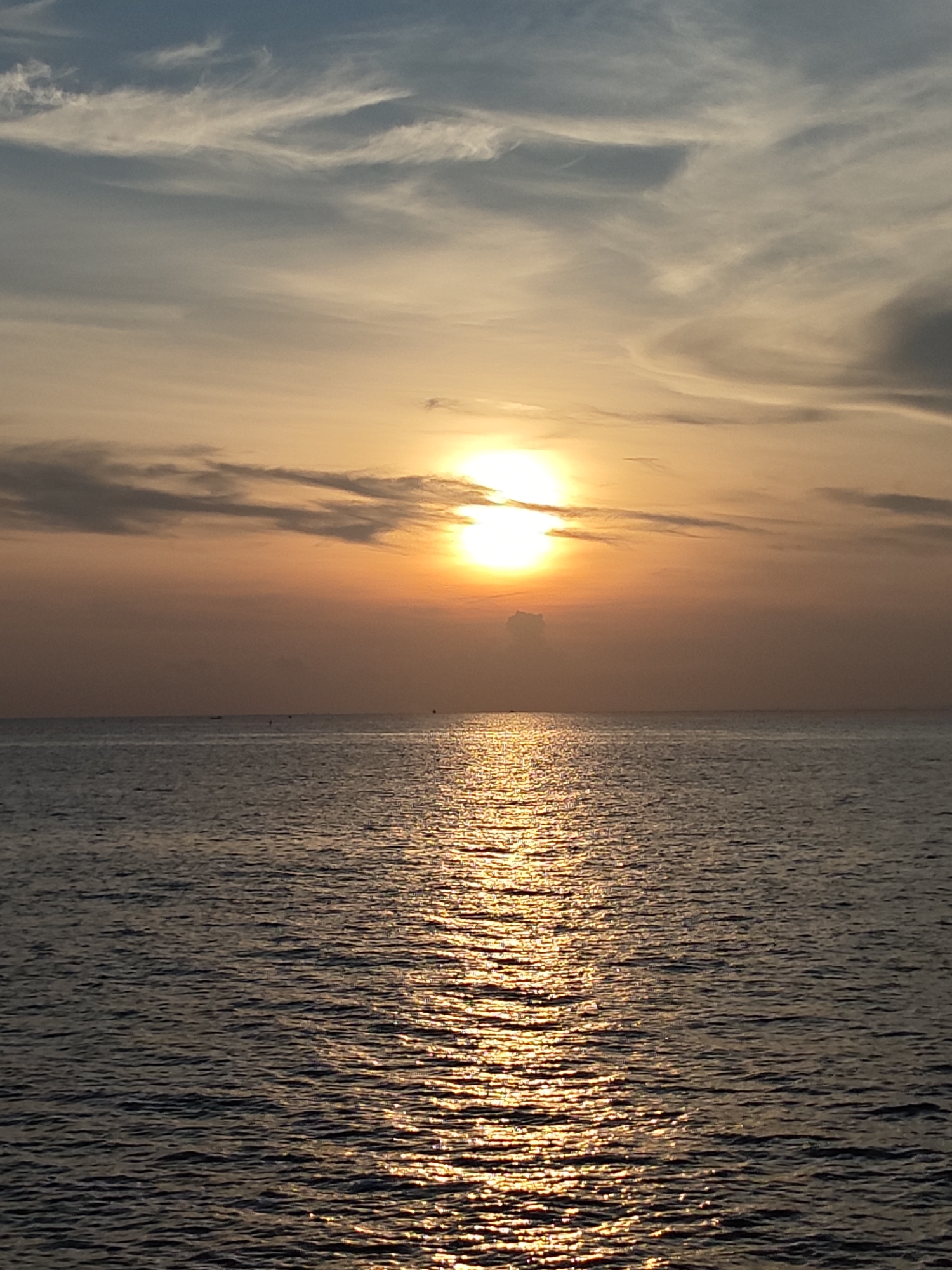 The image captures a sunset over the ocean, likely off the coast of Lhokseumawe, Indonesia, though specific landmarks are not visible. The sun is positioned near the horizon, partially obscured by clouds. The sky is a gradient of colors, transitioning from blues at the top to oranges and yellows around the sun. The water reflects the sunlight, creating a shimmering path towards the viewer. No people are visible, and the scene focuses on the natural environment.