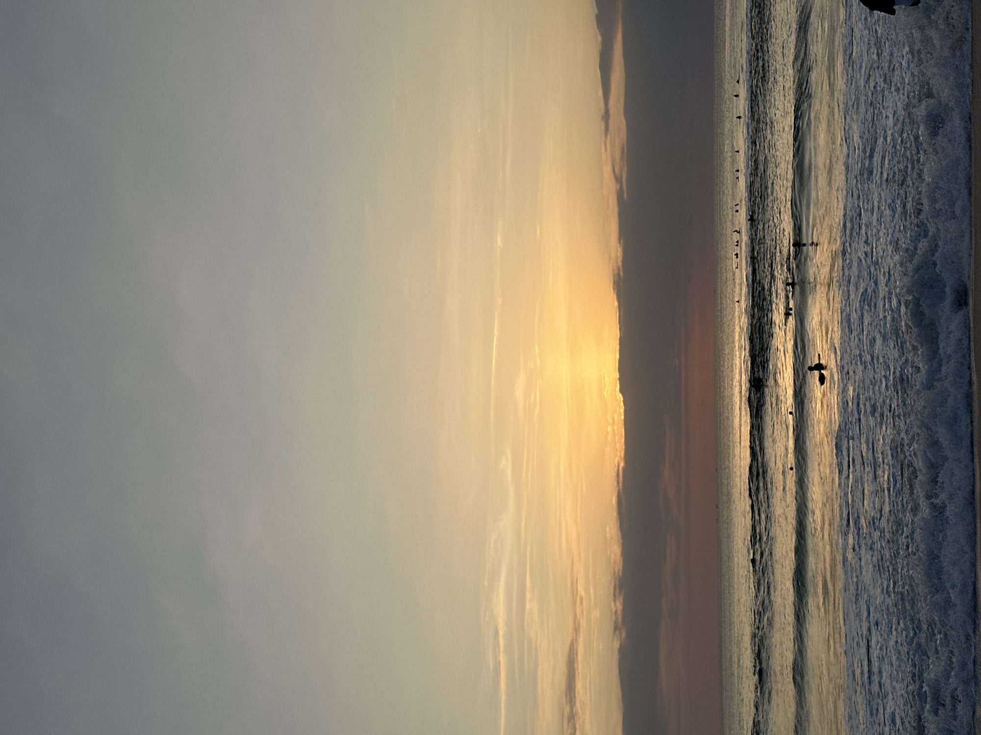 The image shows a sunset over the ocean in Kuta Selatan, Indonesia. The sky is filled with hues of orange, yellow, and grey, with clouds obscuring the setting sun. The ocean is calm with gentle waves breaking on the shore. Several surfers are scattered across the water, some paddling, some waiting for waves. A person is visible on the far right edge of the frame near the shore.