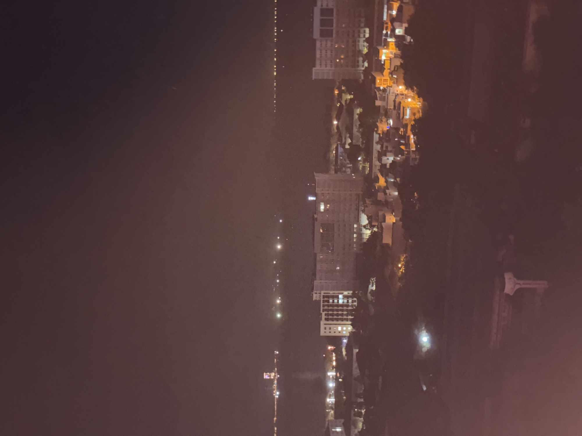 This image is a night-time, aerial view of a coastal city, likely Nha Trang, Vietnam, due to the distant lights appearing to be on a body of water and the architectural style of the buildings.
The scene is dominated by the dark night sky, with a faint haze or mist present. Below the sky, a body of water is visible, lined with numerous distant lights, suggesting a shoreline or a harbor. On the landmass adjacent to the water, several buildings are illuminated.
In the middle ground, there are several multi-story buildings, some appearing to be apartment complexes or hotels, with windows lit up. One prominent white building with visible floors and windows stands out. Other structures, appearing to be lower residential or commercial buildings, are clustered together and lit with warm, orange and yellow lights. Trees are also visible in the foreground and between buildings, appearing as dark masses against the illuminated cityscape.
In the far distance, on the horizon across the water, a single tall, brightly lit structure, possibly a tower or a large building with distinct lighting, is visible. Numerous other faint lights are scattered across the water and along the distant shoreline, indicating the extent of the urban area.
In the immediate foreground, a blurred, dark area is visible, with what appears to be a concrete structure, possibly part of a balcony or a building facade, with a distinct vertical support element. The overall impression is one of a city illuminated at night, viewed from a high vantage point. There are no people or active interactions depicted.