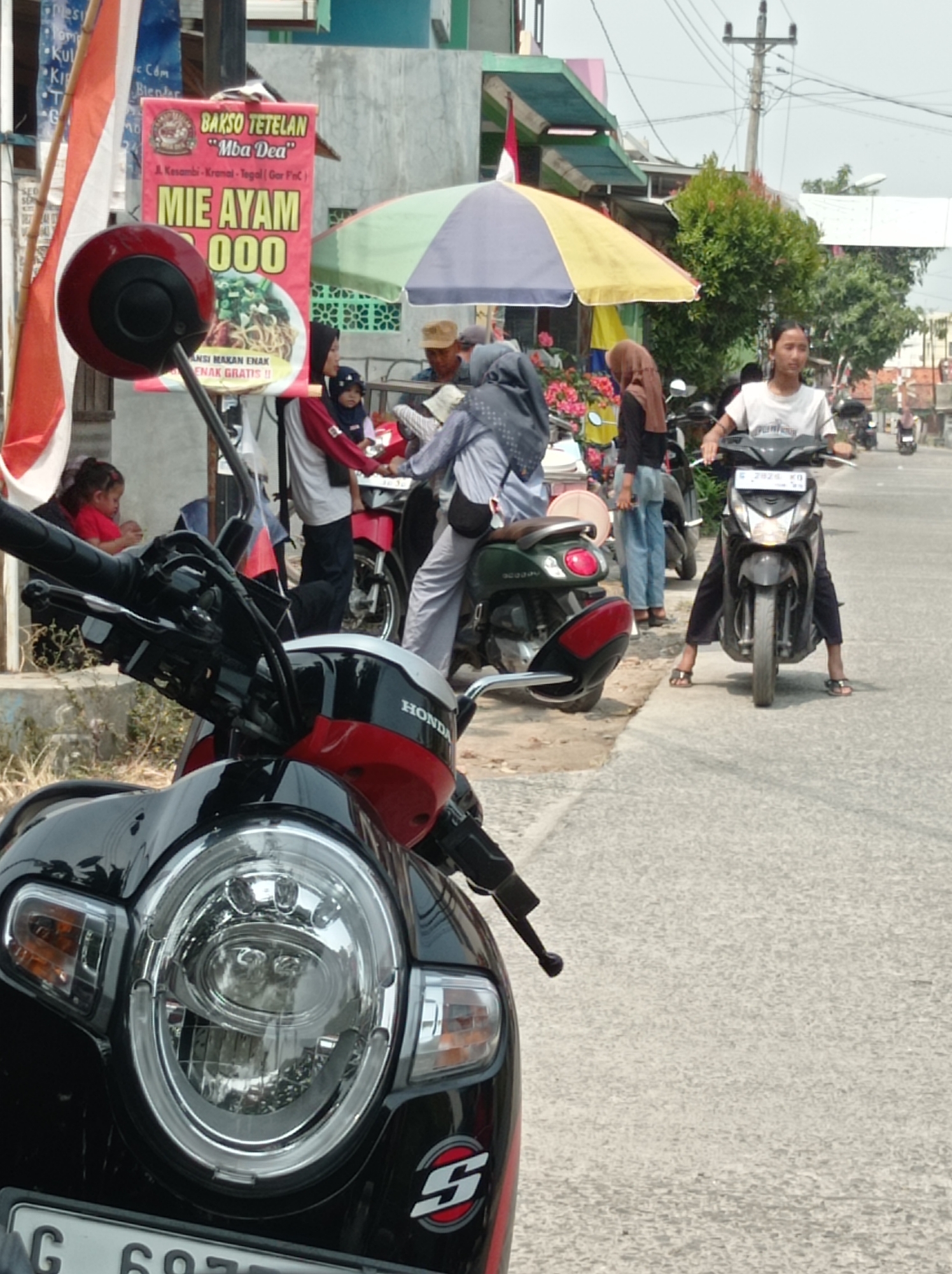 The image depicts a street scene in Kramat, Indonesia, on a bright, sunny day. In the foreground, a black and red scooter with a prominent circular headlight is parked, its handlebars and front fairing clearly visible. Behind the scooter, a busy street is visible with several people and other motorcycles. A young woman wearing a white t-shirt and dark pants is riding a black scooter. Next to her, a person in a grey headscarf and white outfit is holding onto the handlebars of a parked red scooter. Further in the background, a banner advertises "Mie Ayam 000" from "Bakso Tetelan 'Mba Dea'", with text indicating it is located in A. Kasambi-Kramat, Tegal, Gor Foc. Other people are seen in the background, some with headscarves, and more parked motorcycles. The environment includes a sidewalk, a street with asphalt paving, and buildings with awnings and signage. A utility pole with wires is also present. The overall impression is a snapshot of daily life and commerce on a street in Indonesia.