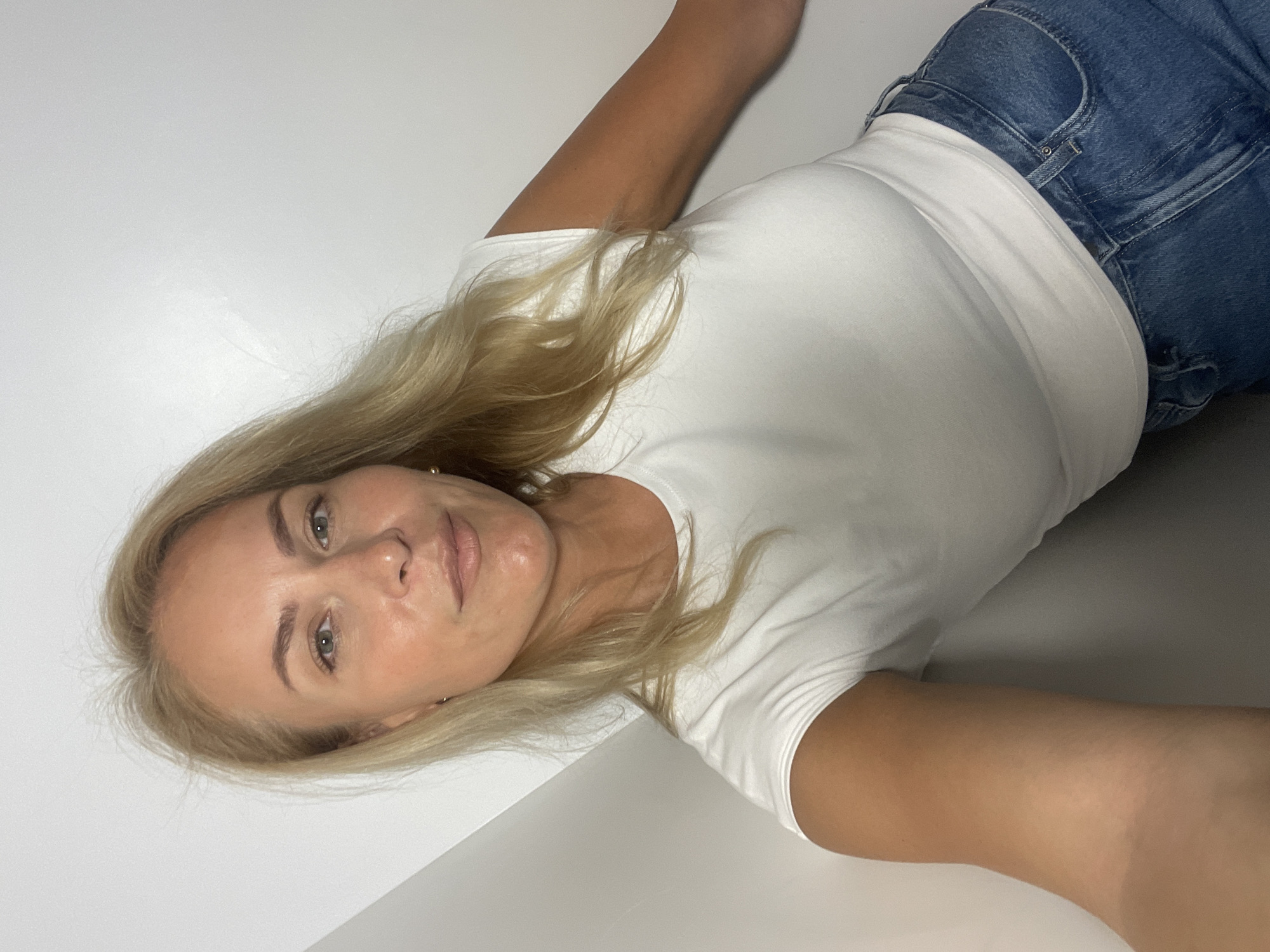 A woman with blond hair is lying on a white surface wearing a white t-shirt and blue jeans. The location cannot be determined from the image.