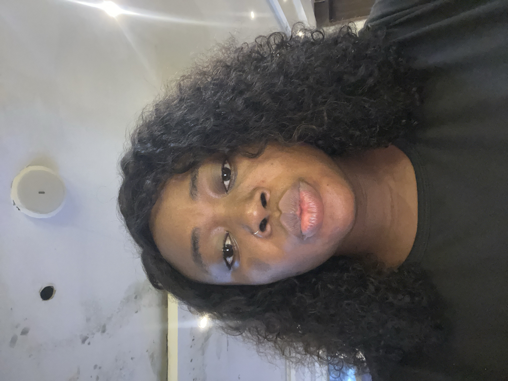 A medium shot shows a Black woman with curly dark hair and a nose ring puckering her lips. She is wearing a black t-shirt. The background is a ceiling with what appear to be lighting fixtures and some water damage. The lighting suggests an indoor setting. There is no visual information that places the scene in Abuja, Nigeria.