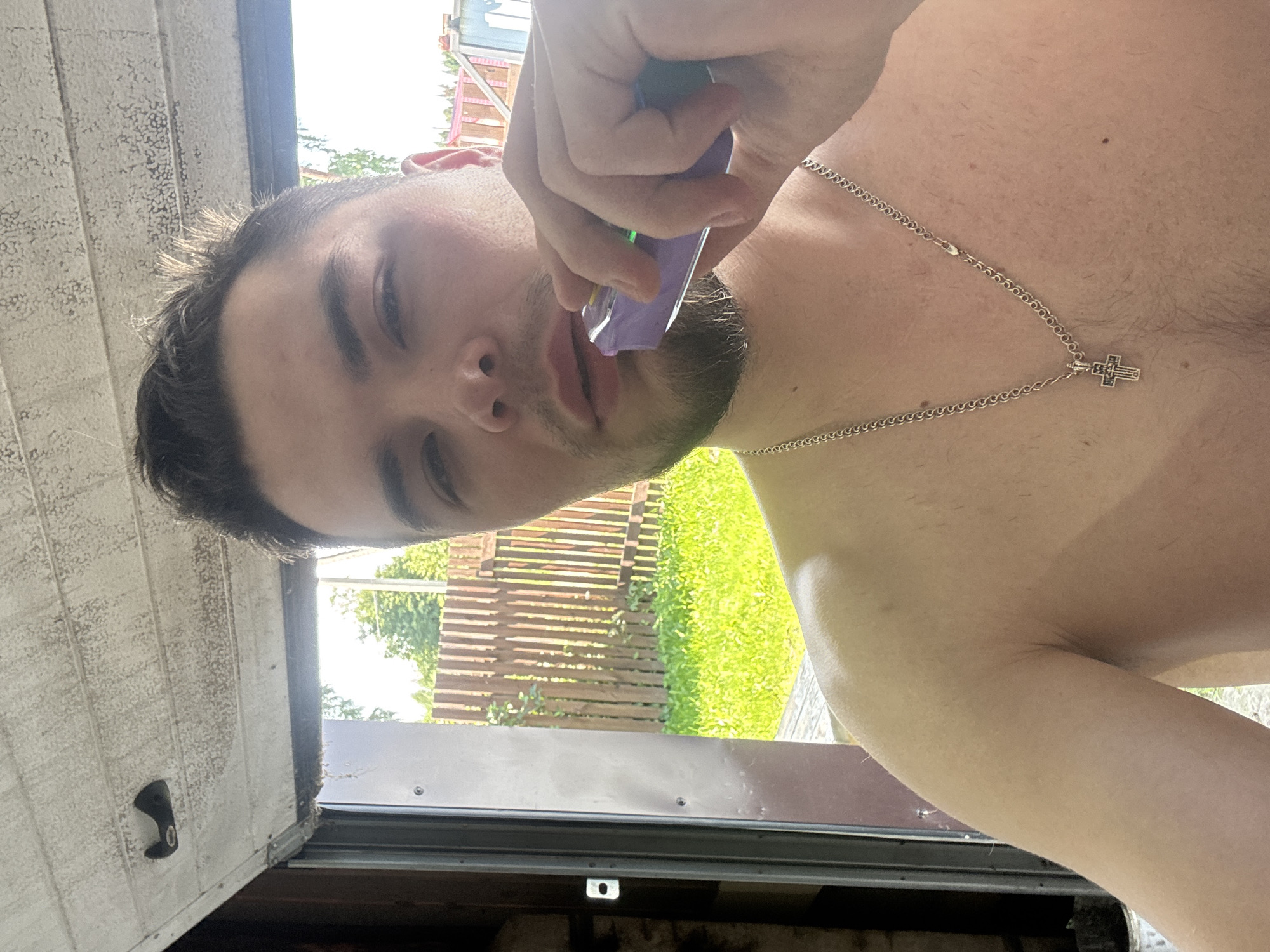 A young man with short, dark hair and a light beard is holding a purple e-cigarette device to his lips, as if about to take a puff. He is shirtless and wearing a silver chain with a cross pendant. He is looking directly at the camera with a neutral expression. The background shows an open garage door, with sunlight illuminating a wooden fence and green grass outside. The man appears to be in a residential area, possibly a backyard, with a wooden deck visible in the foreground. The scene suggests a casual, outdoor setting. The location is identified as Borodulino, Russia, likely a rural or suburban area given the presence of a fence and greenery.