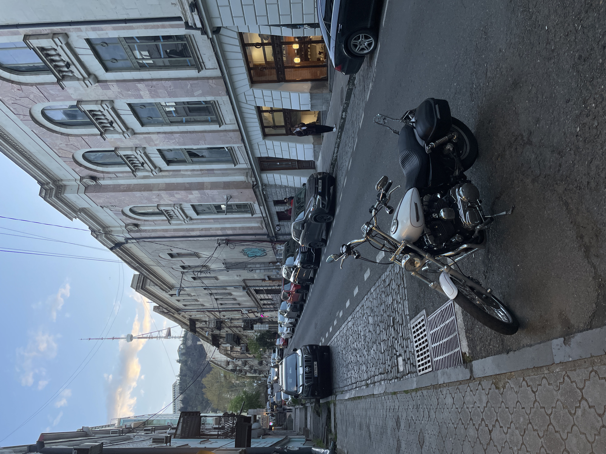 The image presents an urban street scene, likely in Tbilisi, Georgia, with a perspective looking down a street lined with buildings and vehicles.
On the left, a row of multi-story buildings showcases ornate architectural details, including arched windows, decorative ledges, and patterned facades. One building features a brightly lit ground-floor entrance, possibly a shop or restaurant, where a person is seen standing.
The street itself is paved, with a section of cobblestone sidewalk on the right. Numerous cars, including SUVs and sedans, are parked along the street. In the immediate foreground, a white and black motorcycle with chrome accents and a black saddlebag is prominently featured, with another motorcycle partially visible behind it.
In the far distance, against a partly cloudy sky, the distinctive Tbilisi TV Tower rises above a tree-covered hill, serving as a key landmark. Utility lines traverse the upper left portion of the frame.
The lighting suggests it is late afternoon or early evening, with soft, ambient light casting gentle shadows. The weather appears mild with some clouds in the sky.
A notable visible detail is a Georgian license plate, "KK 790 KK," on a black SUV parked further down the street. The scene depicts a typical city environment with stationary vehicles and people casually present.