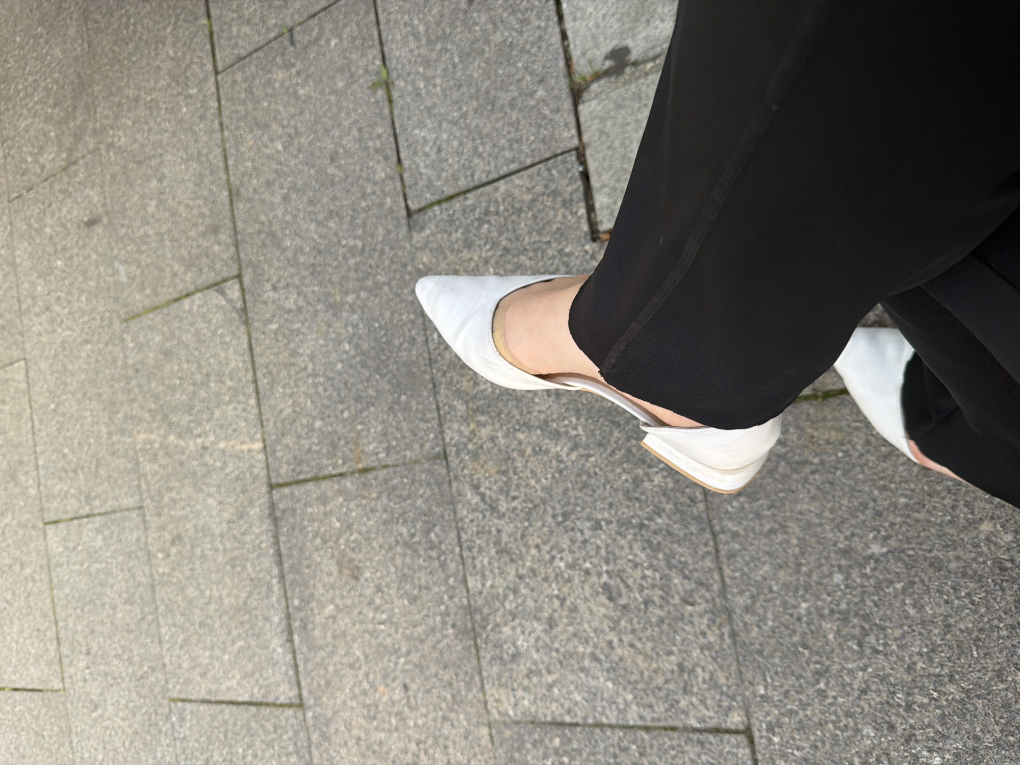 An overhead shot shows a person's legs crossed, sitting on a textured gray paved surface. The person is wearing black pants and white pointed-toe flats. The shoes have a low heel and appear to be made of a matte material, possibly leather or a synthetic alternative. One shoe is more visible, showing the pointed toe and the heel strap. The pants are a solid black and have a visible seam running down the side. The paved surface consists of rectangular stone tiles with visible grout lines. The lighting suggests it is daytime, possibly outdoors.