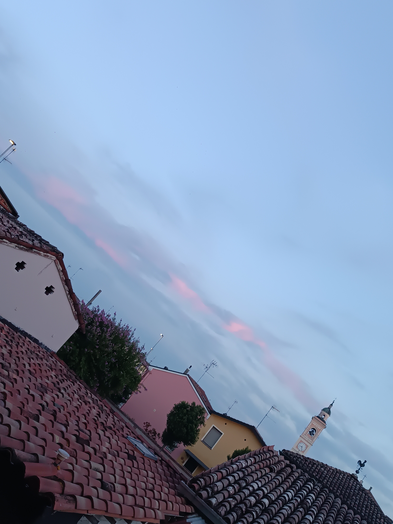 The image captures a picturesque twilight scene in Viadana, Italy, characterized by a soft, pale blue sky streaked with delicate pink clouds. The perspective is tilted, offering a view from a rooftop overlooking a charming Italian town. In the foreground, terracotta roof tiles create a textured pattern, with glimpses of neighboring buildings with white and pink facades. A prominent feature is the distant church tower, its clock face visible and its spire reaching towards the heavens, suggesting the heart of the town. Lush green trees and flowering bushes add a touch of nature to the urban landscape. The overall atmosphere is serene and tranquil, hinting at the peaceful ambiance of Viadana at dusk.