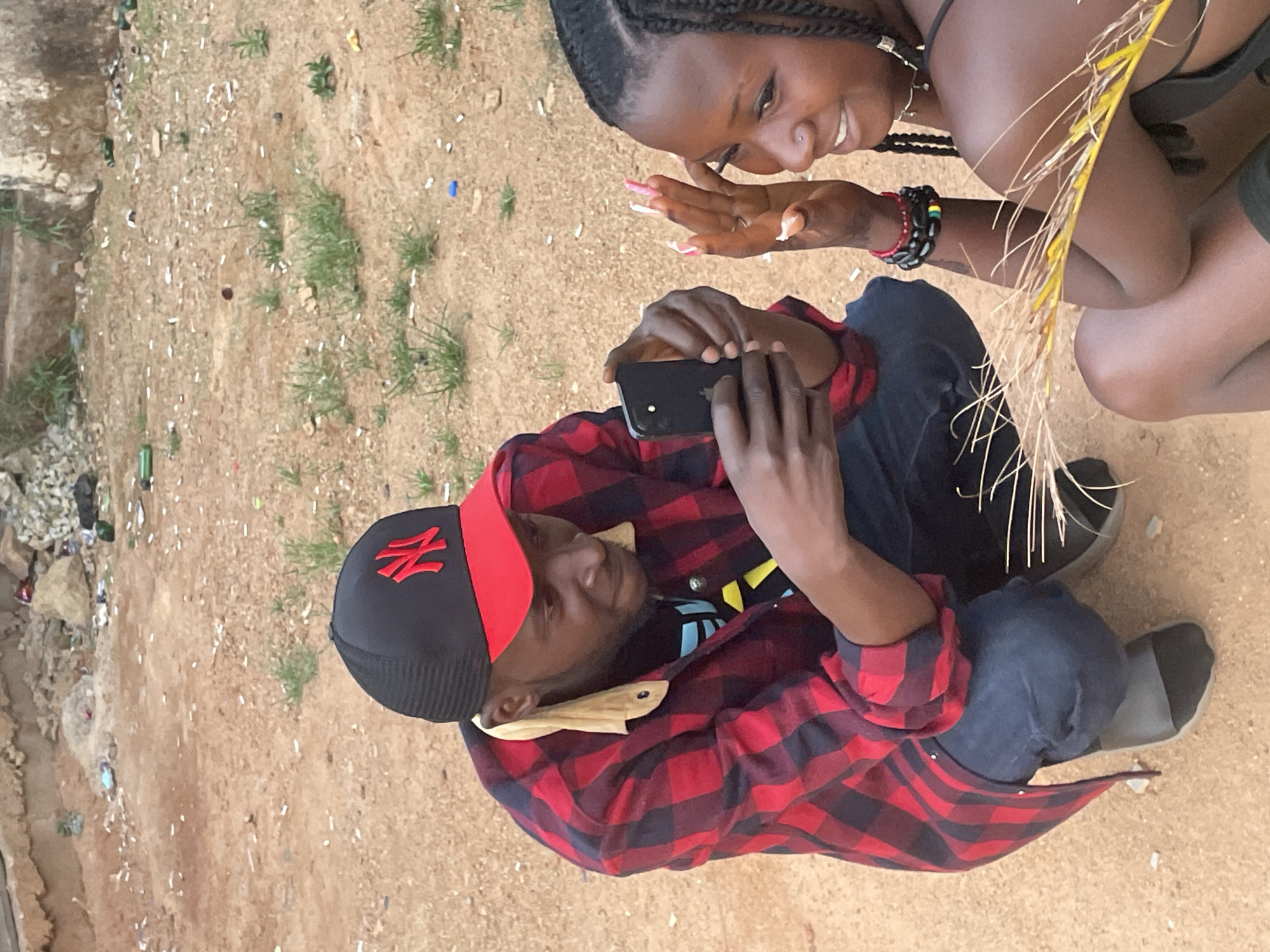 Captured outdoors in Barakin Akawo, Nigeria, the scene presents a man and woman interacting. The man, wearing a black and red NY baseball cap and a red and black plaid shirt, is seated and holding a smartphone, seemingly taking a photo or video. A woman with braided hair, adorned with a bracelet on her wrist, is positioned next to him, looking at the phone screen. They are seated on sandy ground scattered with trash and sparse greenery.
